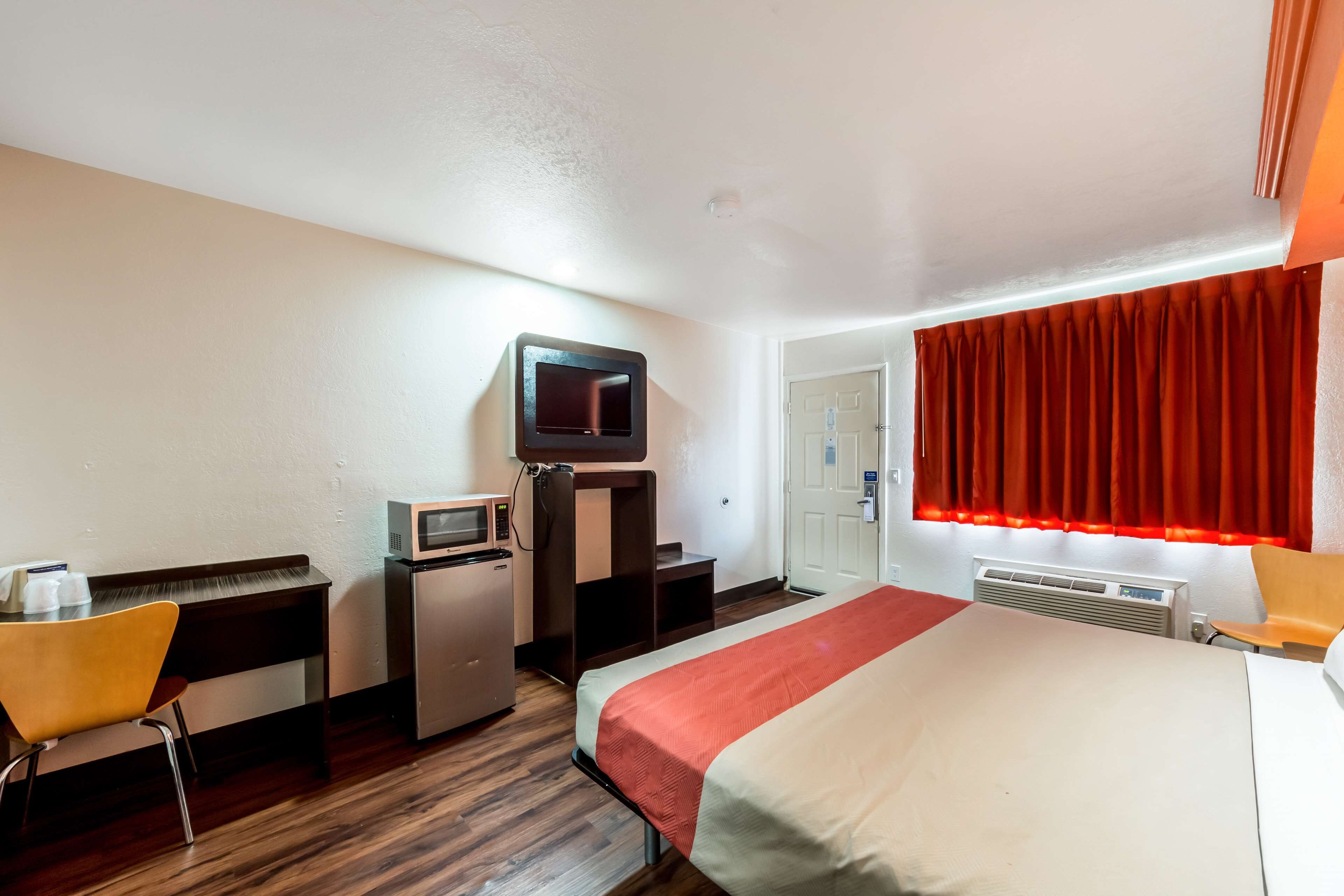 A hotel room with a bed, a desk with a computer, a microwave, a TV, and a red curtain.