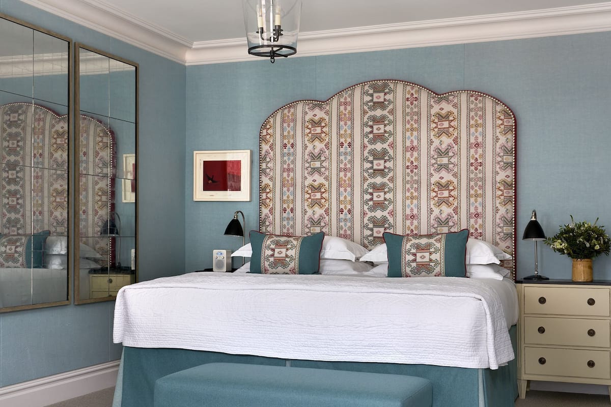 A bedroom with a large bed, a bench, a dresser, a mirror, and a wall with patterned wallpaper.