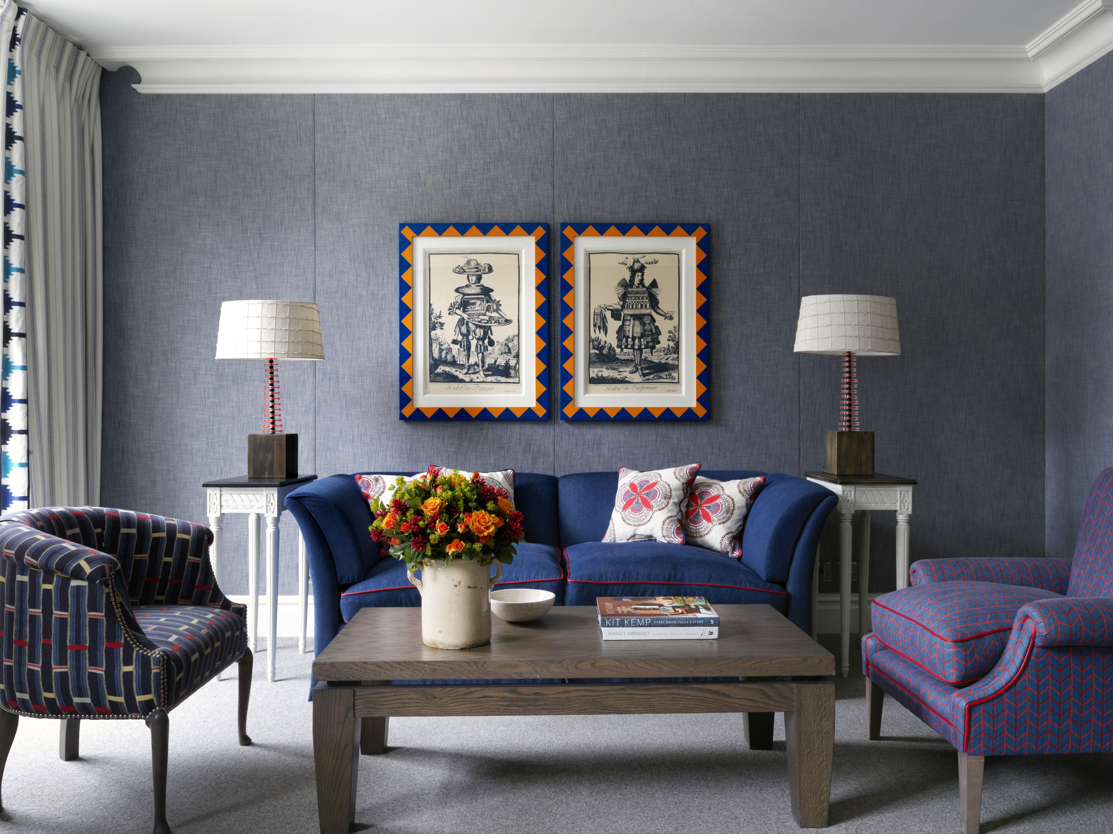 Una sala de estar con un sofá azul, sillones estampados, una mesa de centro y dos cuadros en la pared.