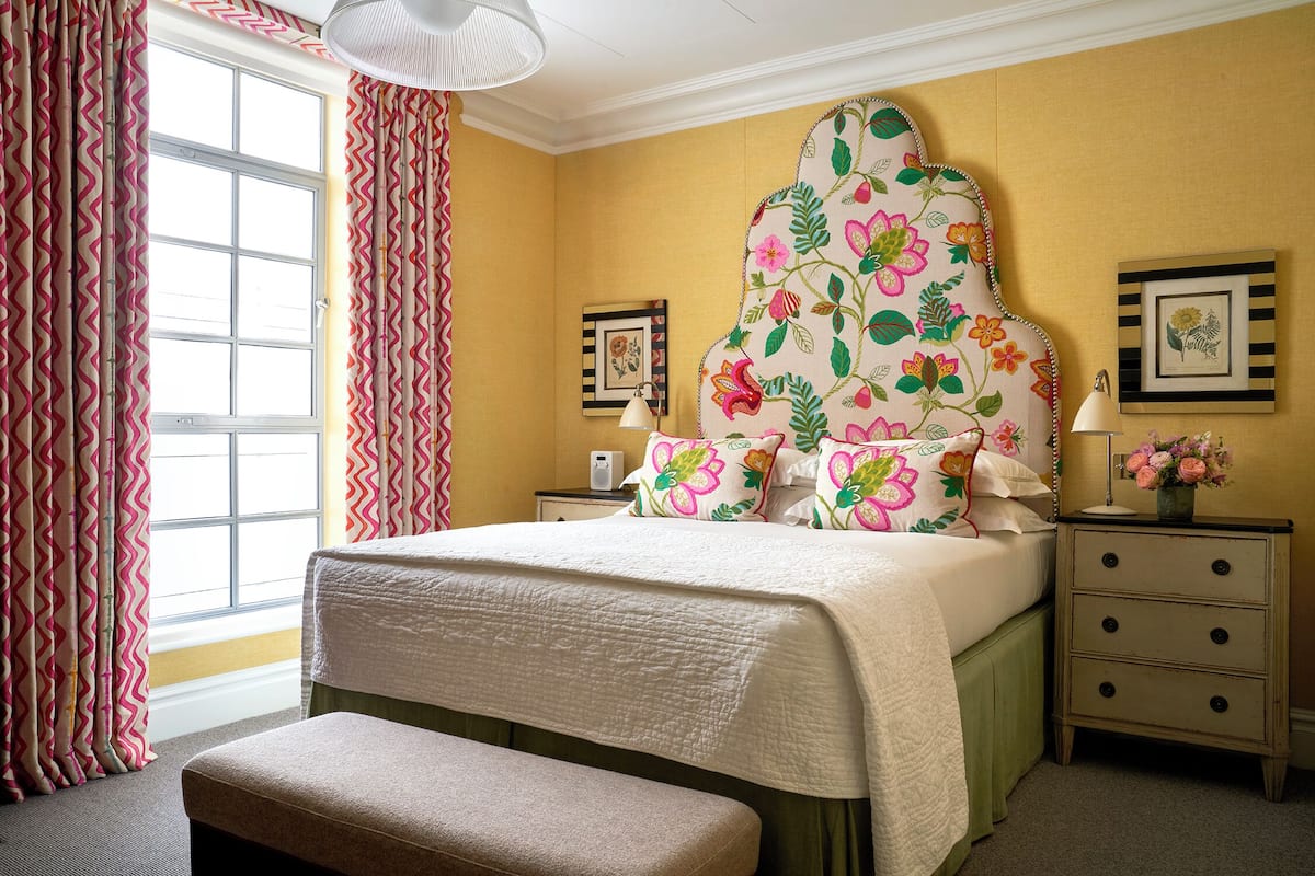 A neatly made bed with a floral headboard, a bedside table with a lamp, and a picture frame on the wall.