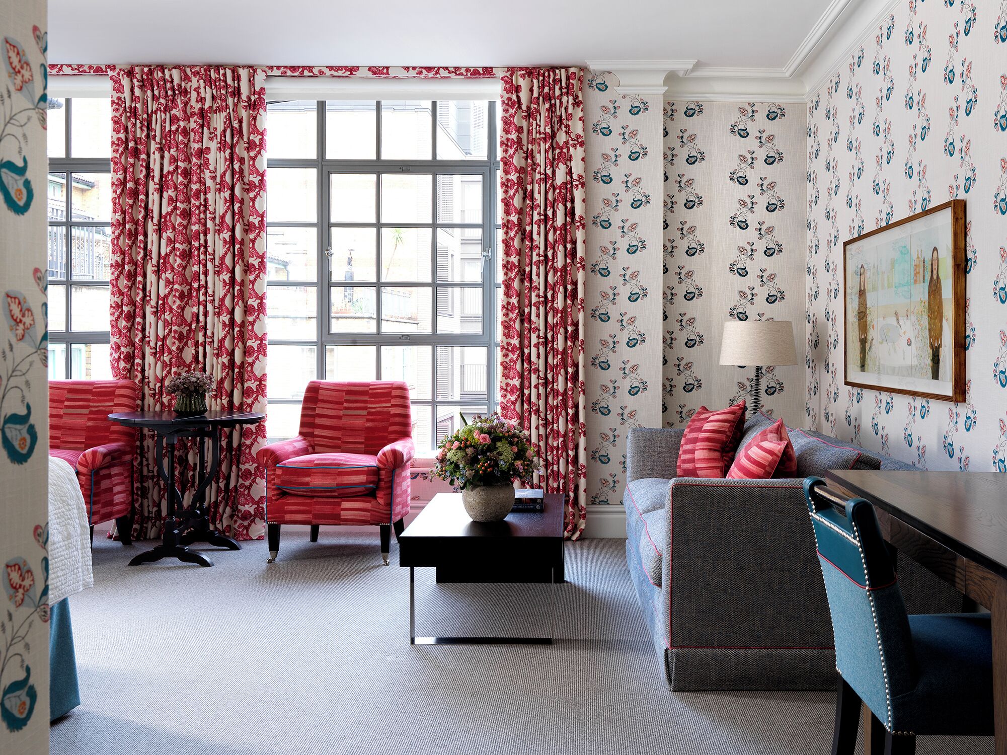 A room with a large window, red curtains, a sofa, a chair, a small table with a vase, and patterned wallpaper.