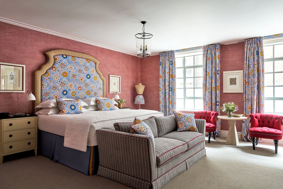 A bedroom with a large bed, a sofa, a red armchair, a small table, and a window with curtains.
