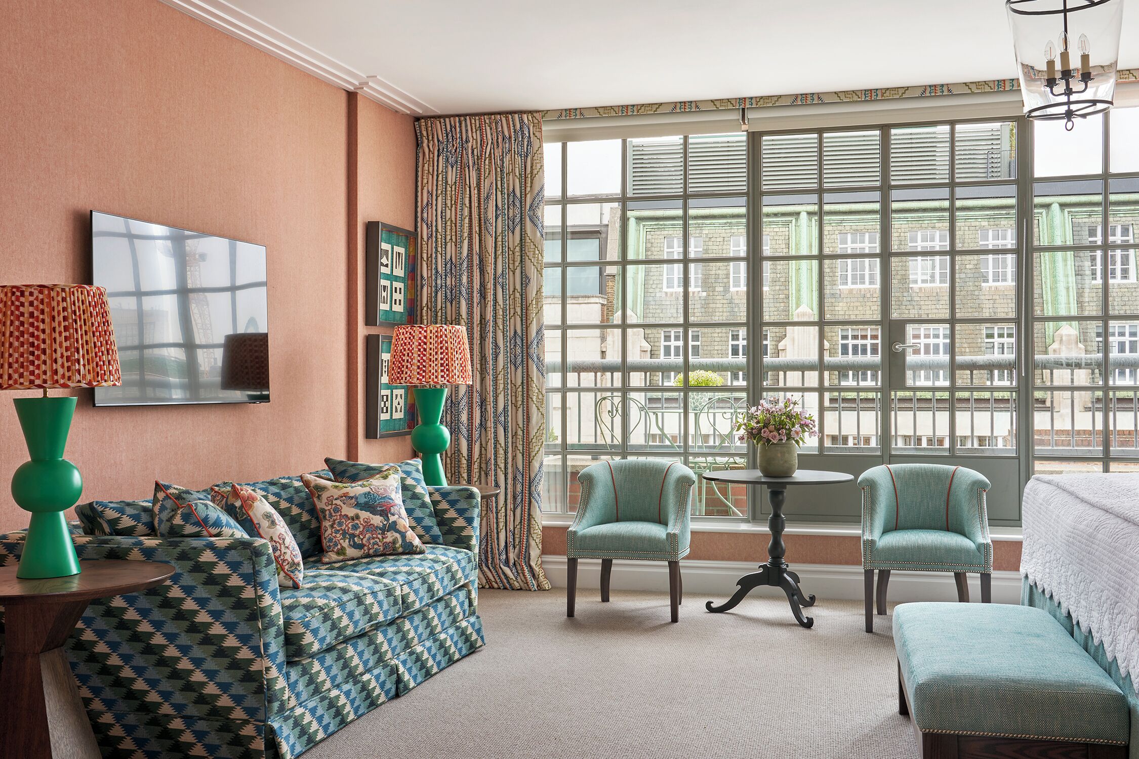 A room with a patterned sofa, two armchairs, a small table with a vase, and a large window with a view of buildings.
