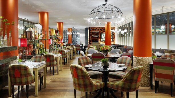 Un restaurant doté de nombreuses tables et chaises, de grandes colonnes orange et d’une décoration variée comprenant des plantes et un grand lustre.