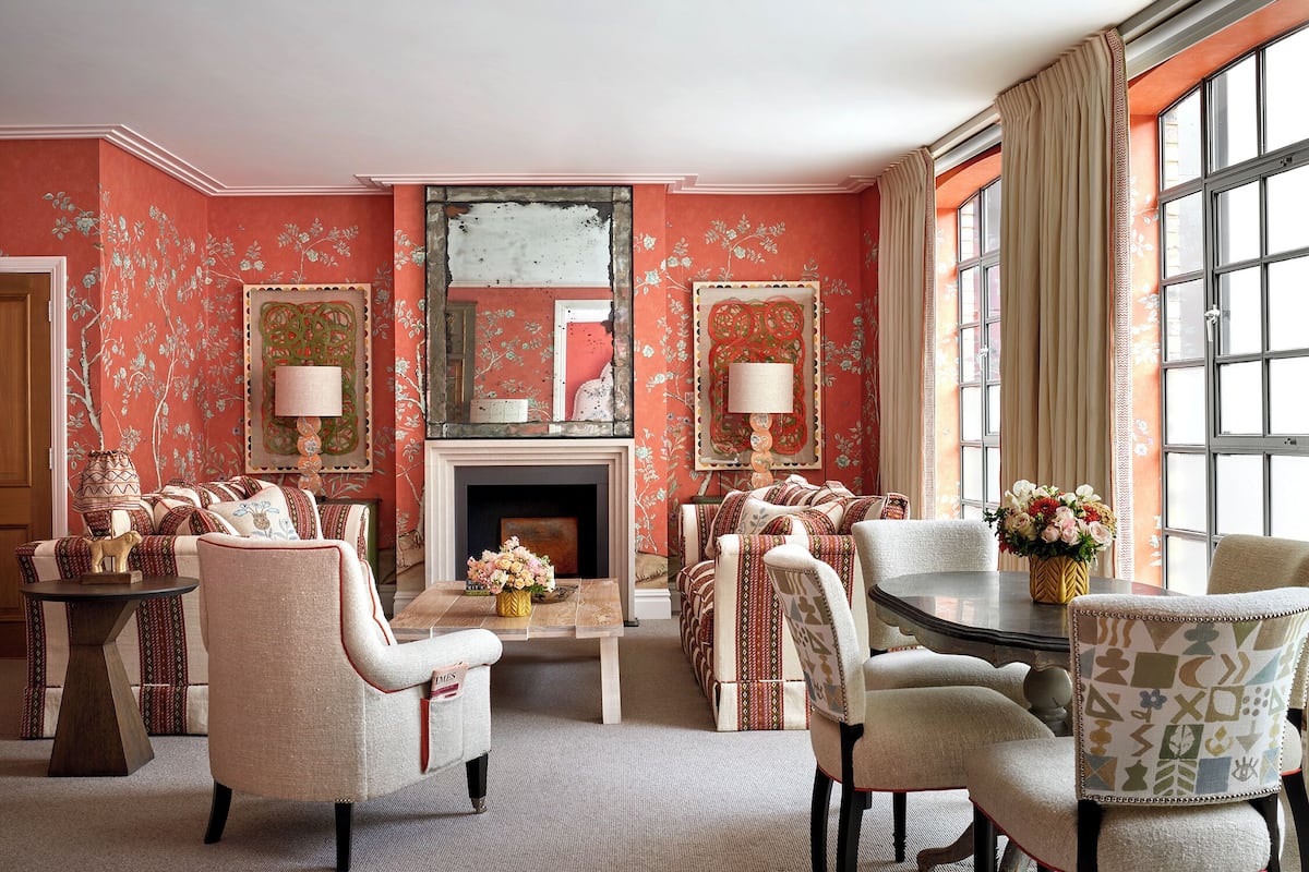 A living room with a fireplace, floral wallpaper, and a large window with curtains.