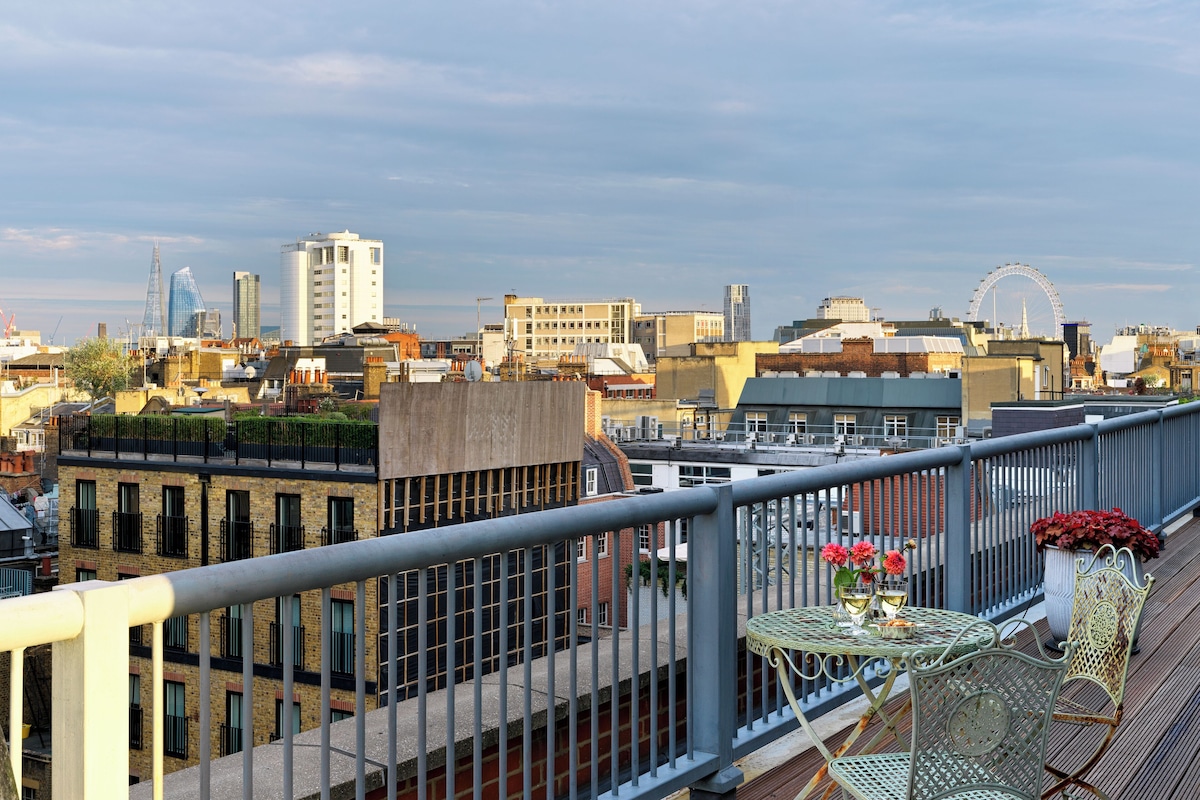A rooftop terrace with a metal railing, a table with chairs, and a view of the cityscape including buildings and a Ferris wheel.
