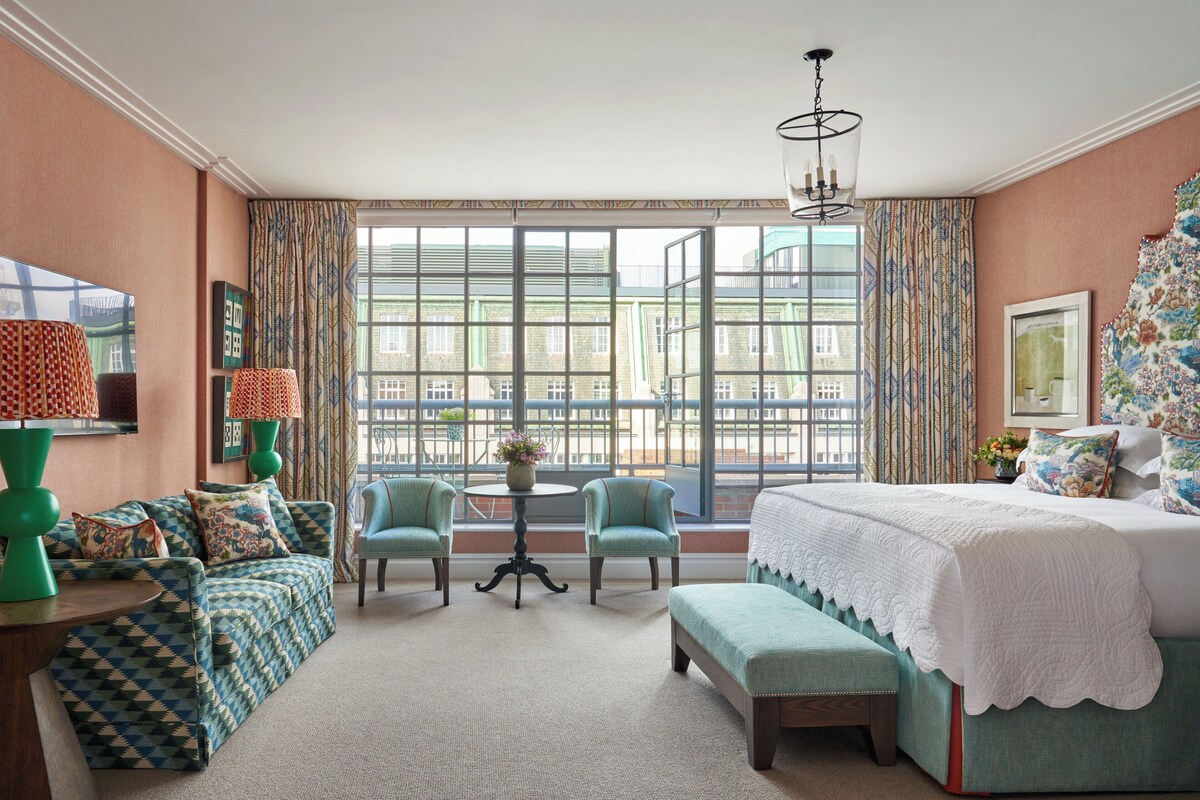 A bedroom with a large window, a bed, a sofa, two chairs, a small table, and a bench.