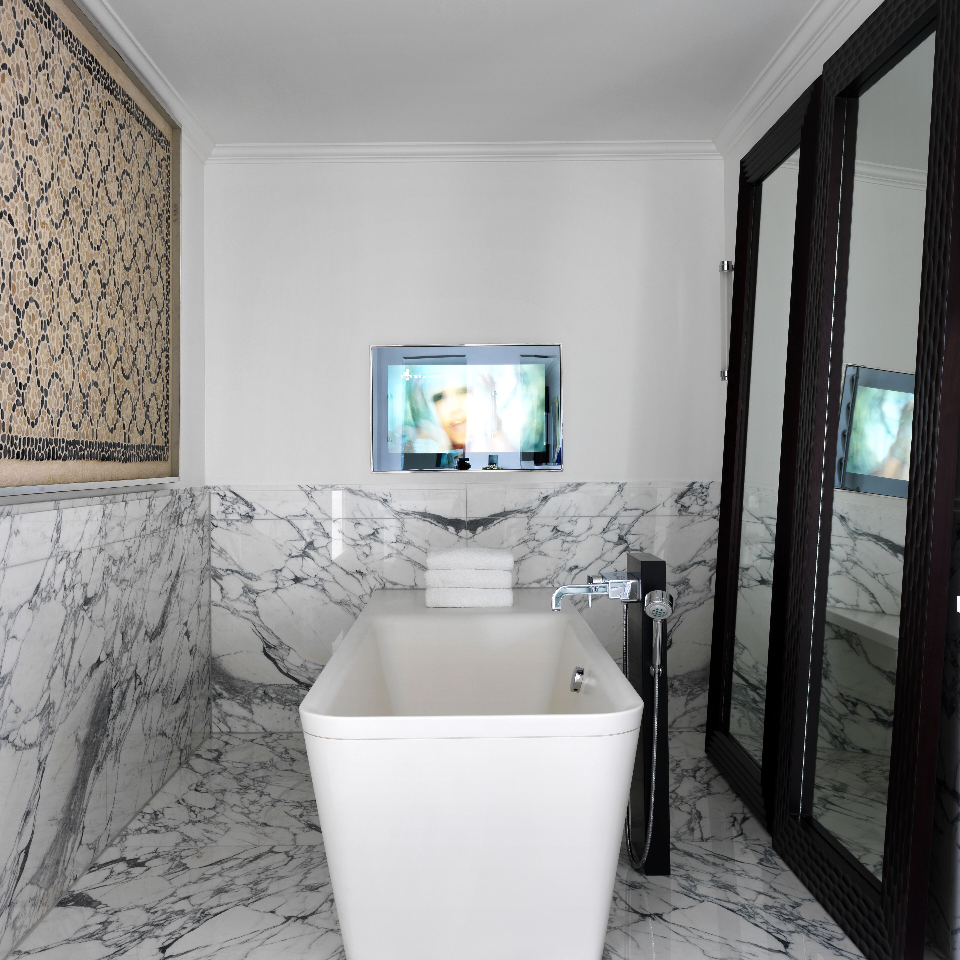 A modern bathroom with a marble floor, a white bathtub, a handheld showerhead, and a television mounted on the wall.