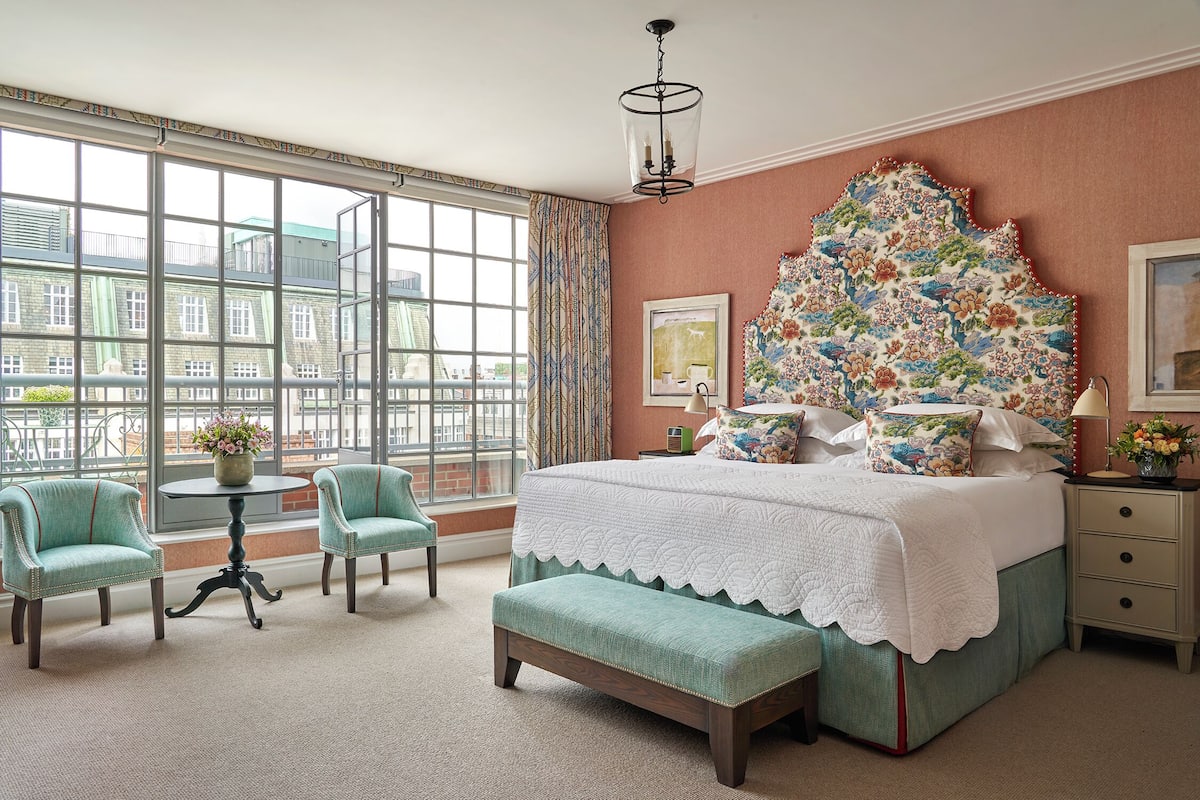 A bedroom with a large window, a bed with a floral headboard, two chairs, a small table, and a bench.