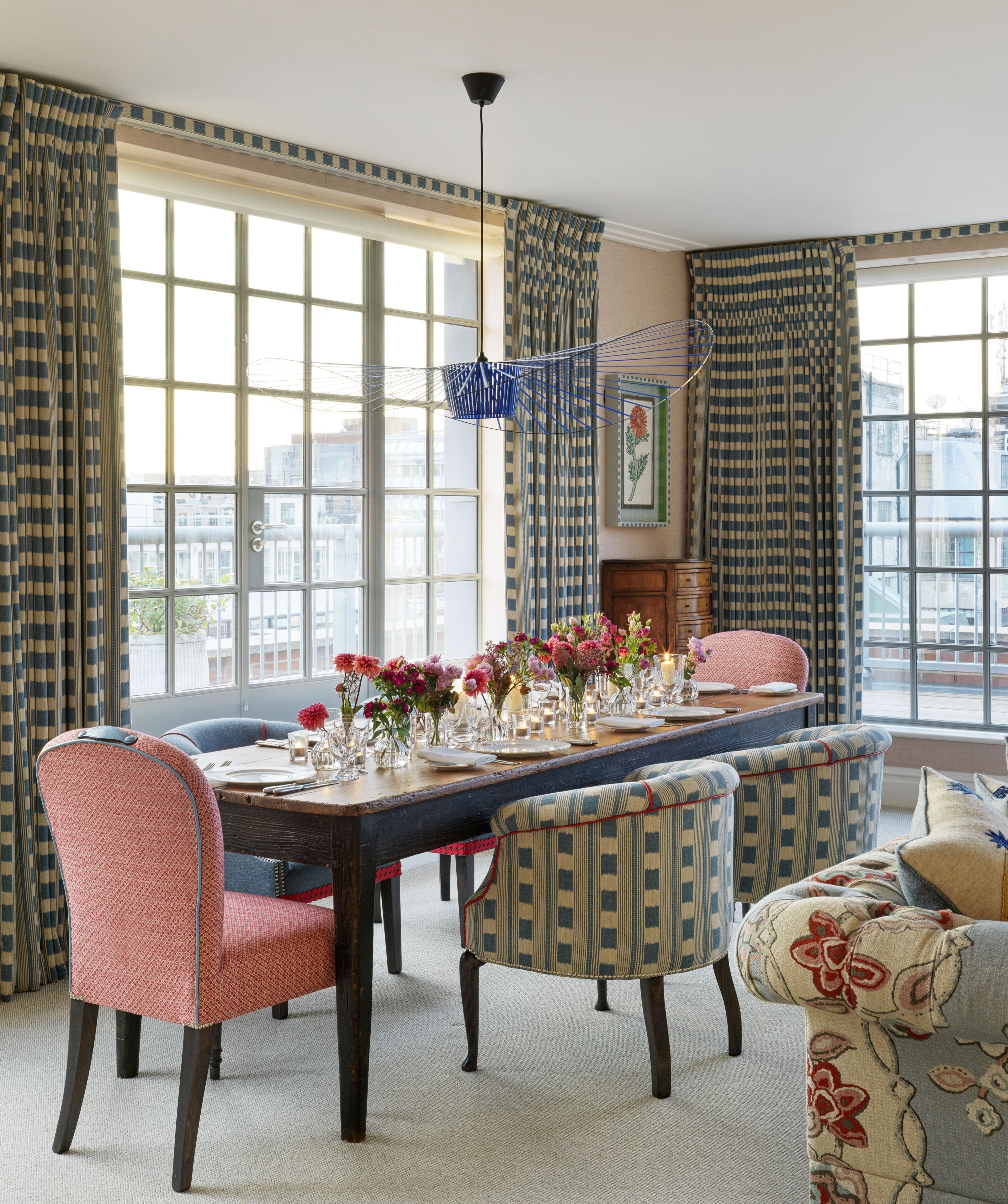 A dining room with a long table set for six, surrounded by patterned chairs, large windows with curtains, and a hanging light fixture.