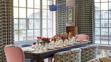 A dining room with a long table set for six, surrounded by patterned chairs, large windows with curtains, and a hanging light fixture.