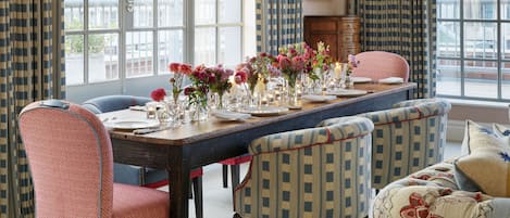 A dining room with a long table set for six, surrounded by patterned chairs, large windows with curtains, and a hanging light fixture.
