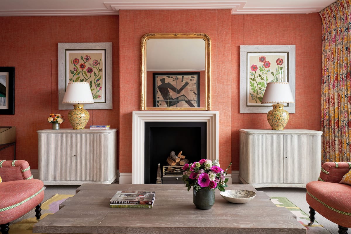 A living room with a fireplace, two armchairs, a coffee table with books, and floral vases.