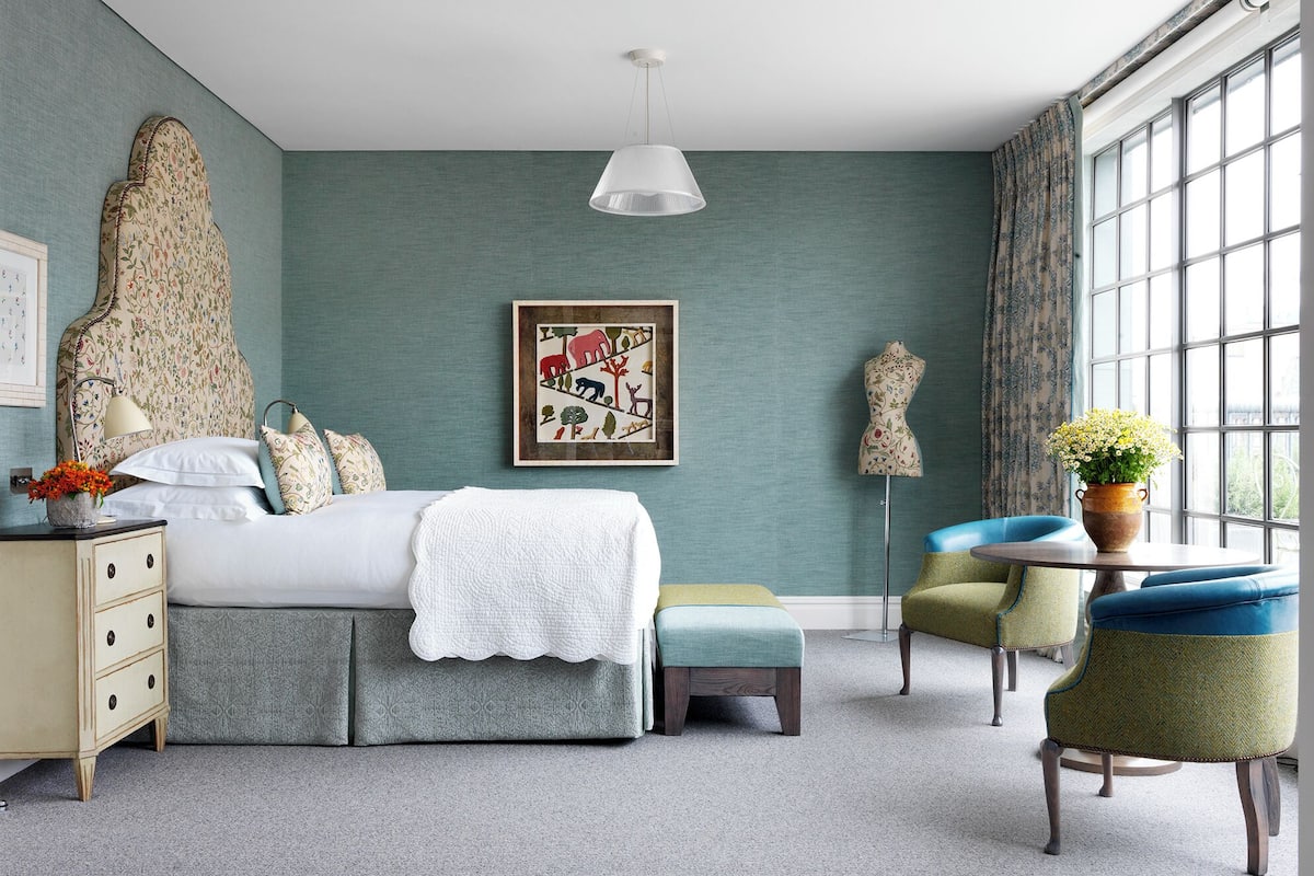 A bedroom with a large bed, a bedside table with a vase of flowers, a chair, and a window with curtains.