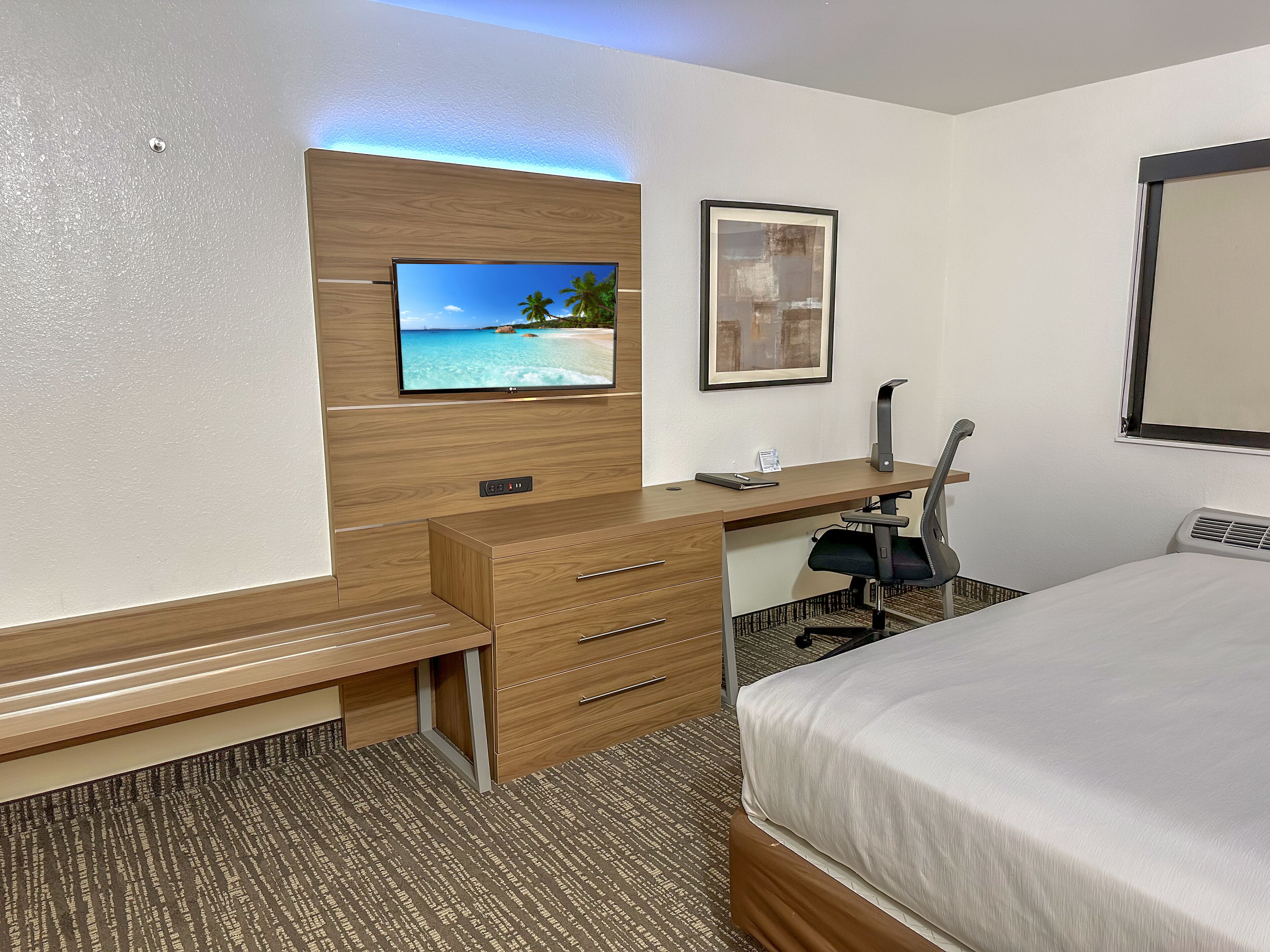 Hotelkamer met een bed, een bureau met lades, een stoel, een aan de muur bevestigde televisie en een ingelijst schilderij.