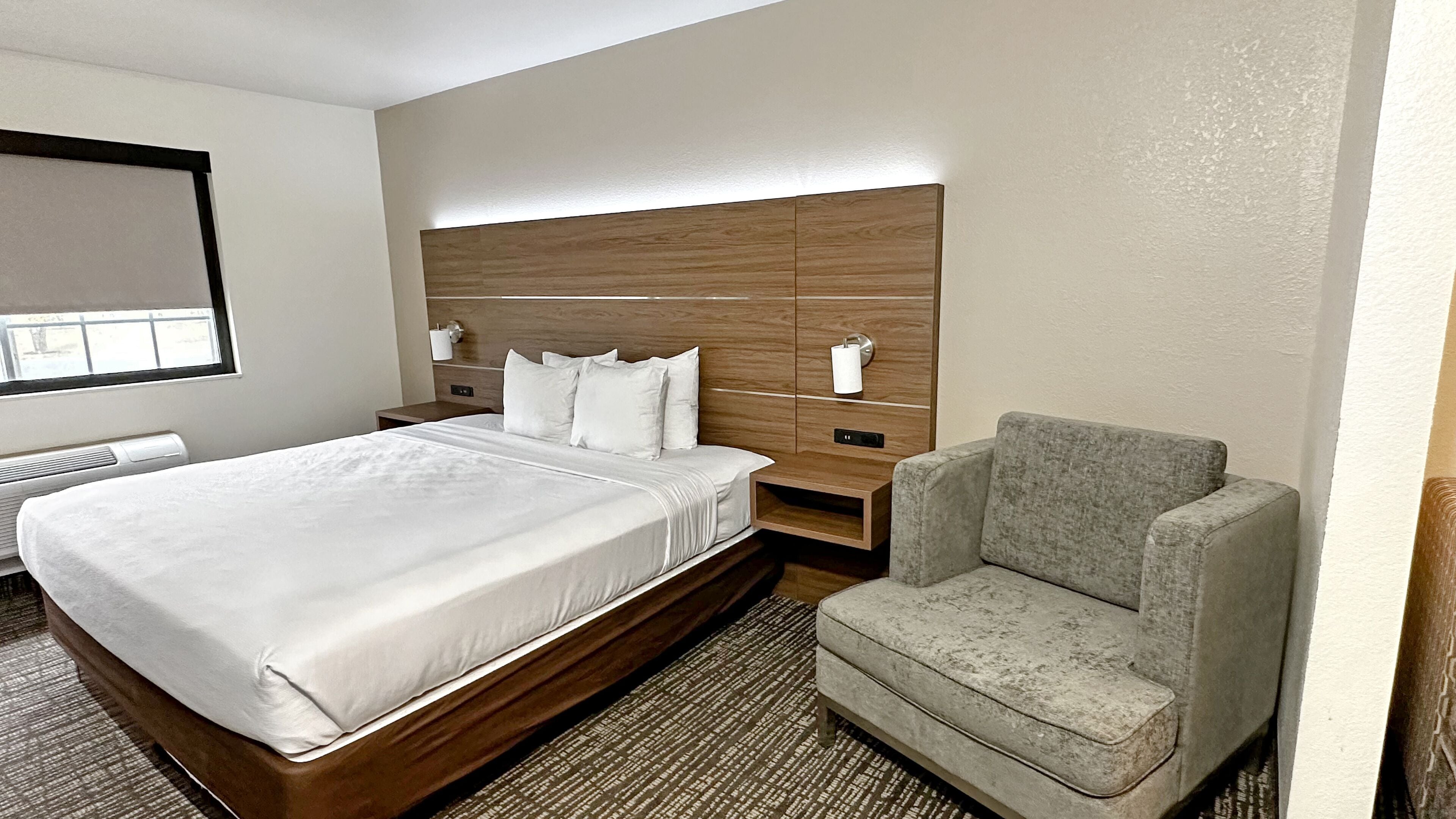 Une chambre d’hôtel avec un grand lit, un fauteuil gris, une tête de lit en bois et une fenêtre avec des stores.