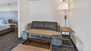 A compact hotel room with a sofa bed, a small wooden table, a bedside table with a lamp, and a headboard.