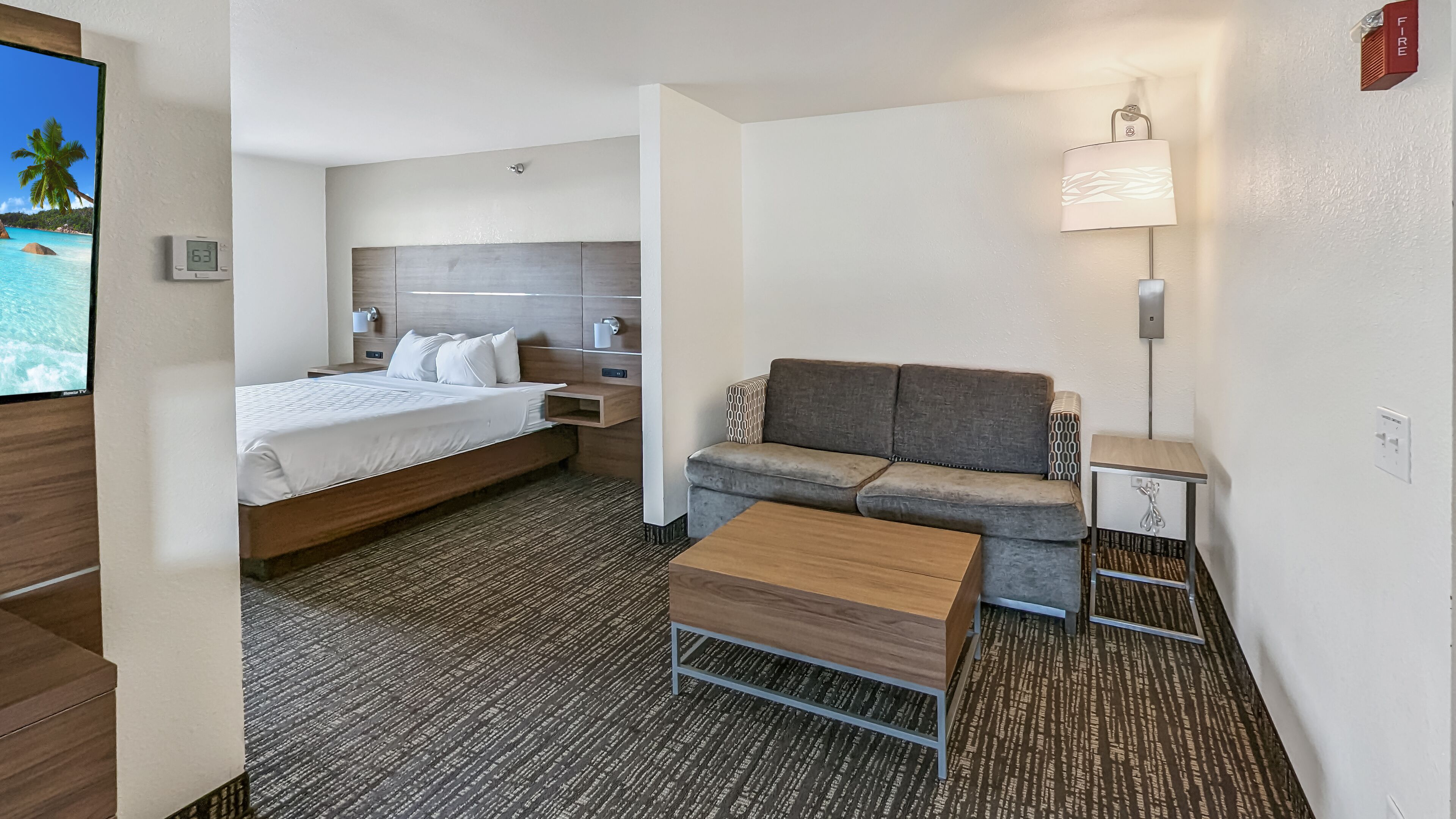 A hotel room with a bed, a sofa, a coffee table, a TV, a lamp, and a fire alarm.