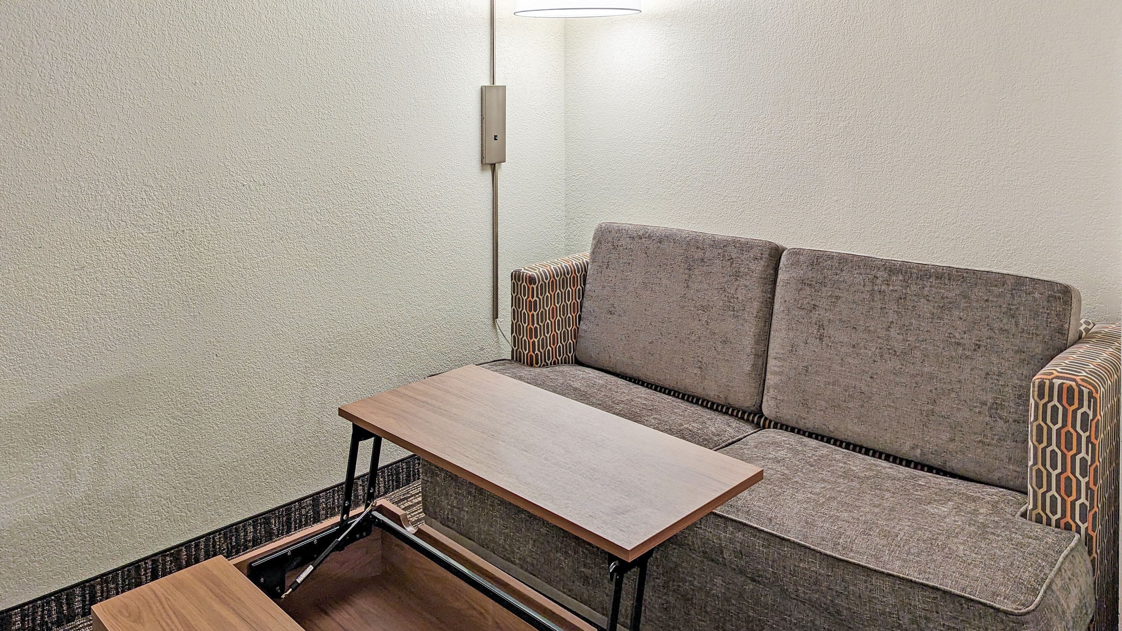 Quarto pequeno e compacto com um sofá, uma mesa de madeira e um ponto de luz fixado na parede.
