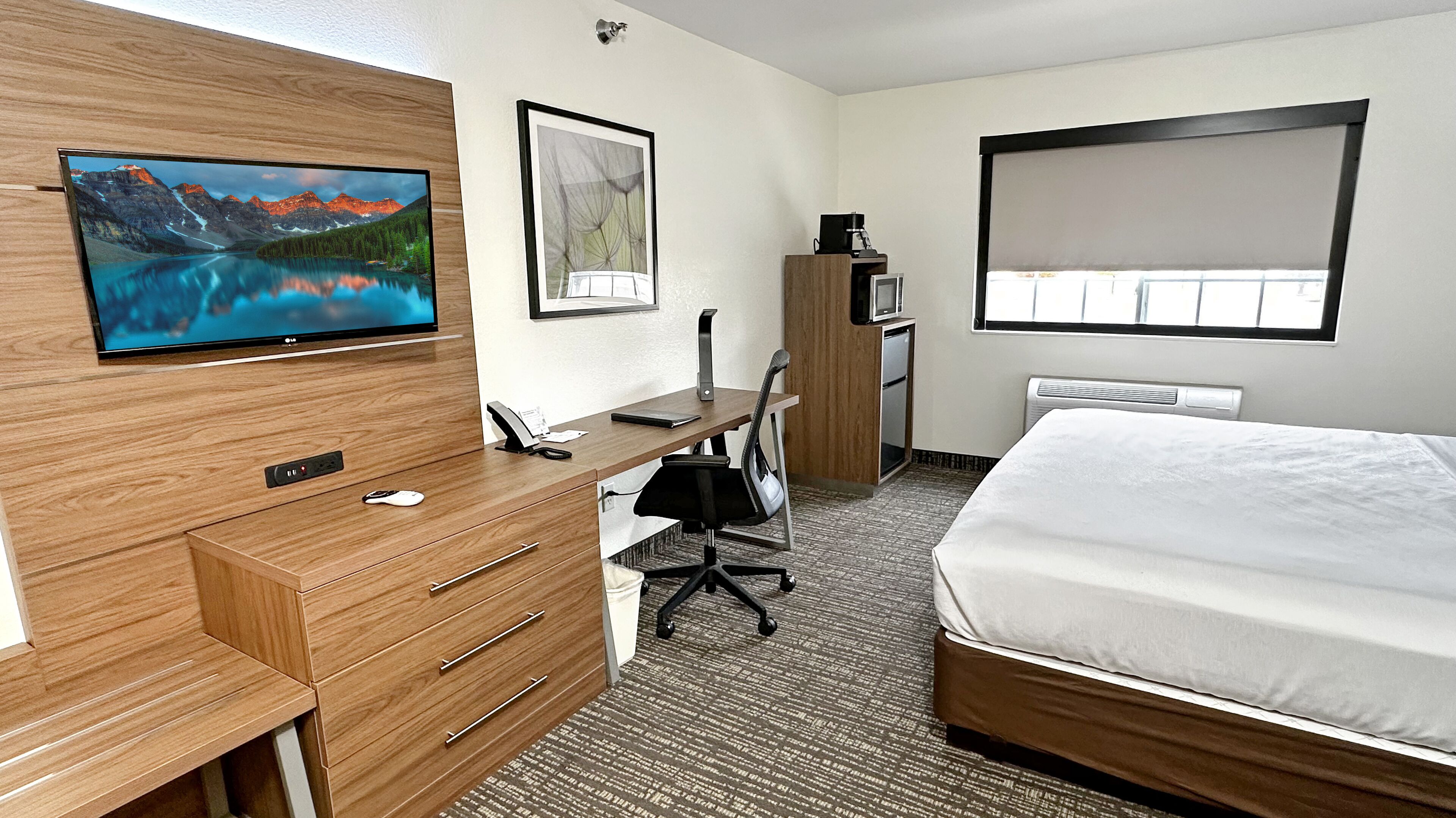 A hotel room with a bed, a desk with a chair, a television mounted on the wall, and a framed picture.