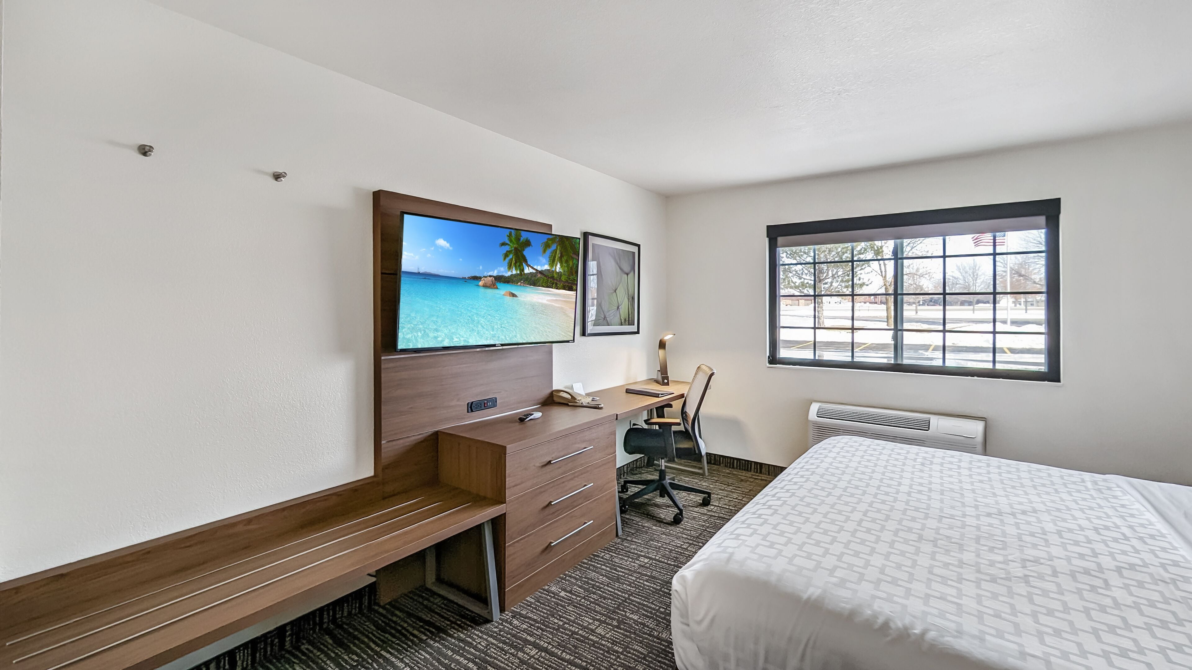 Habitación de hotel con cama, escritorio con silla, televisión en la pared y ventana con vista a árboles y un estacionamiento.