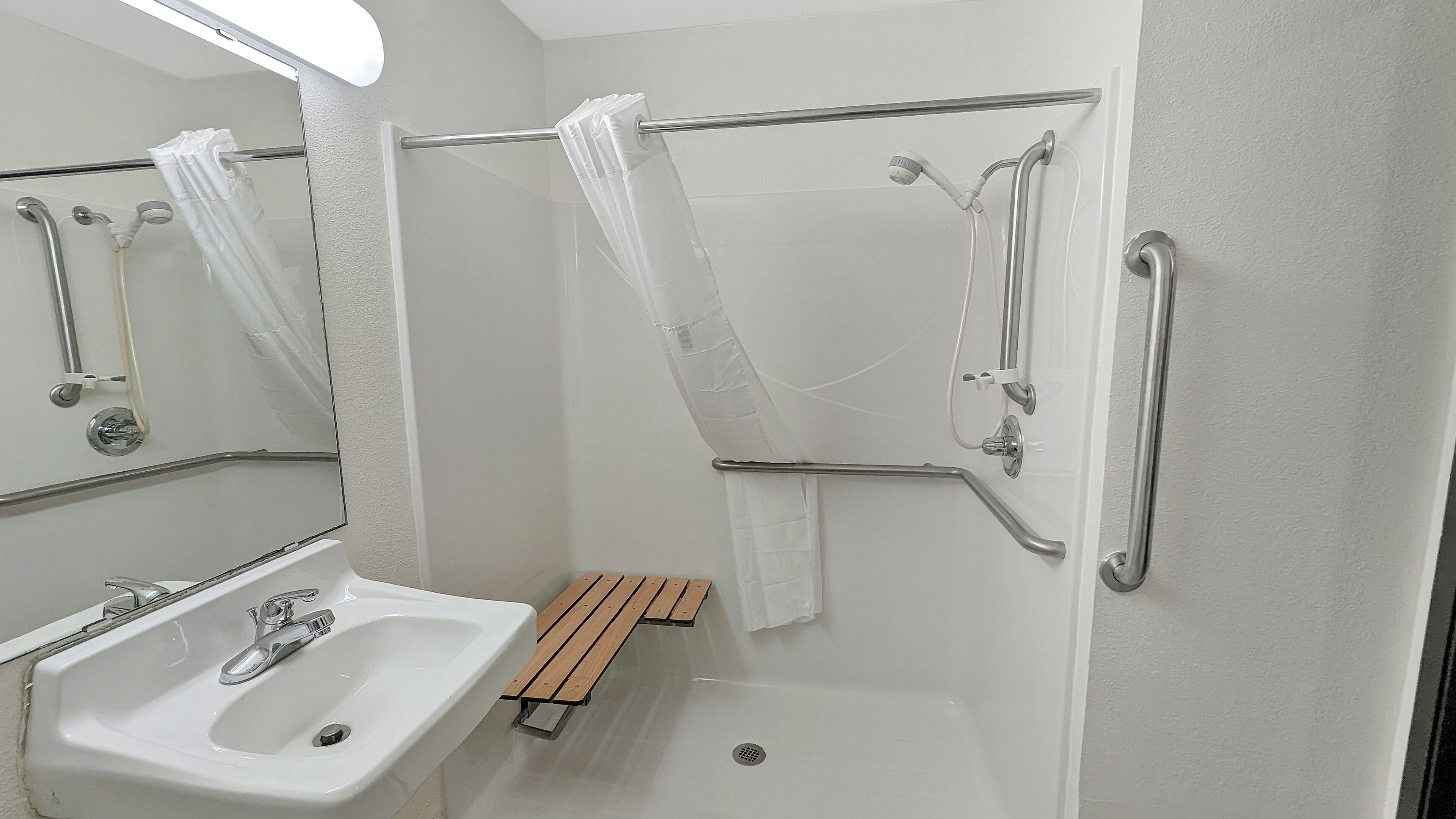 A bathroom with a white sink, a mirror, a shower with grab bars, and a wooden bench.