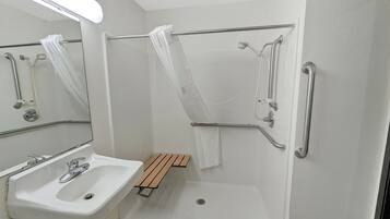 A bathroom with a white sink, a mirror, a shower with grab bars, and a wooden bench.