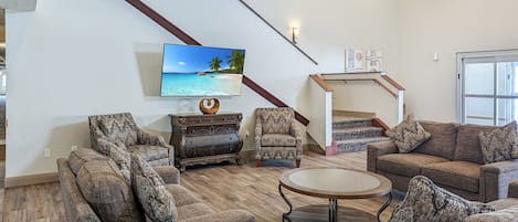 Een woonkamer met gestreepte banken, een salontafel, een aan de muur bevestigde televisie, een trap met uitzicht en een kroonluchter.