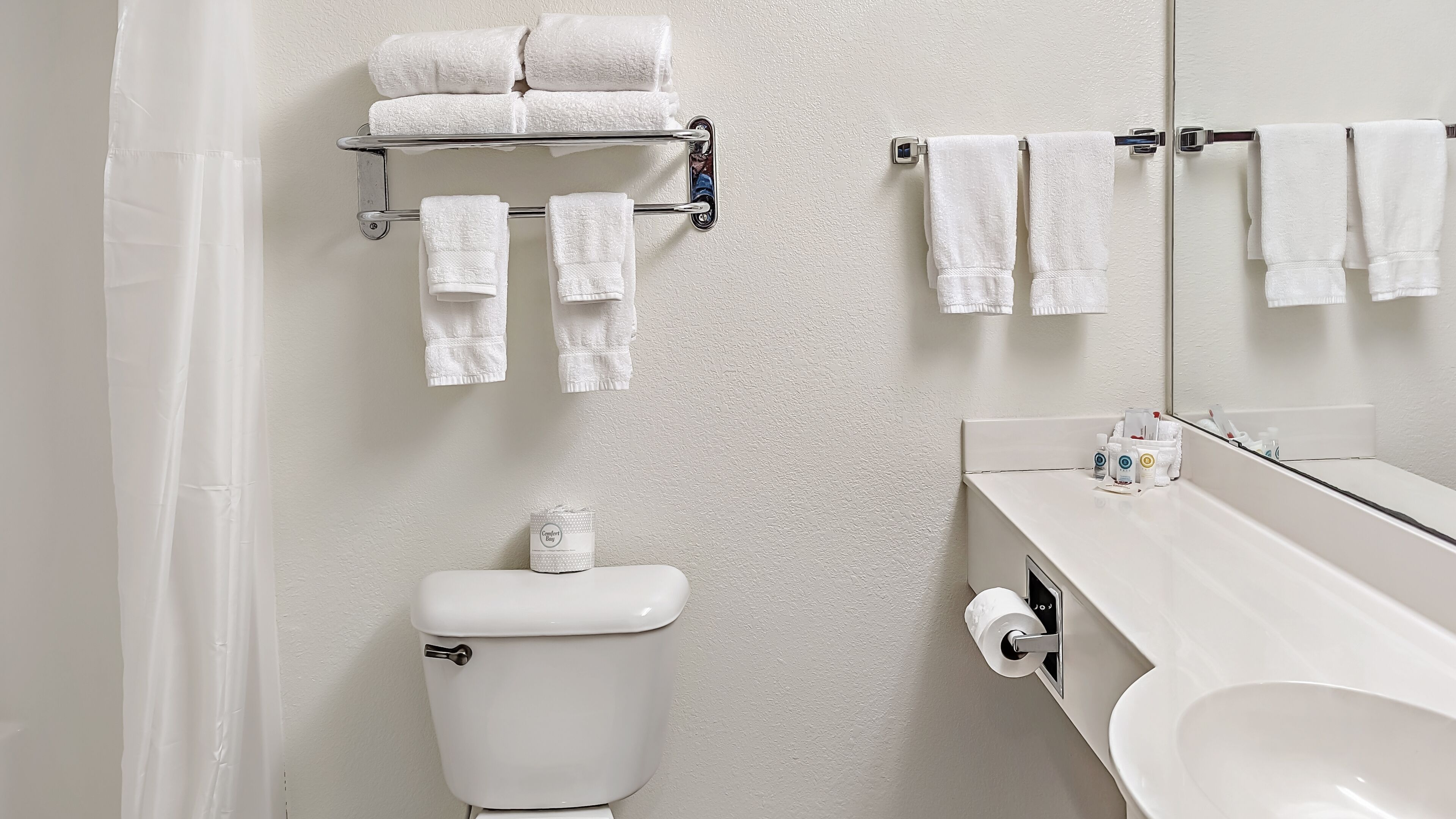 A bathroom with a toilet, a towel rack, a shelf with towels, a sink, and a mirror.