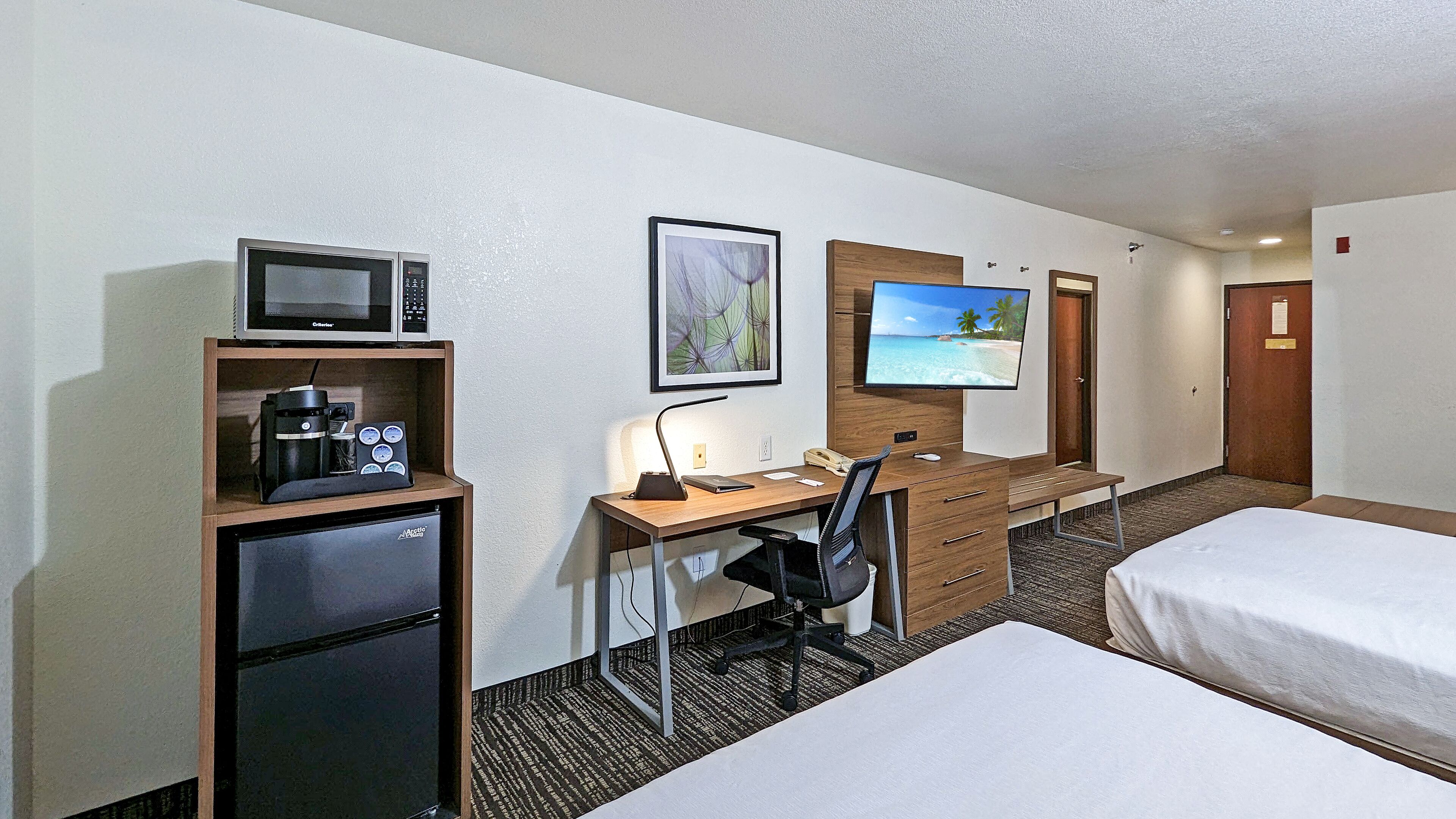 Quarto de hotel com cama, escrivaninha com computador, cafeteira, micro-ondas e televisão.