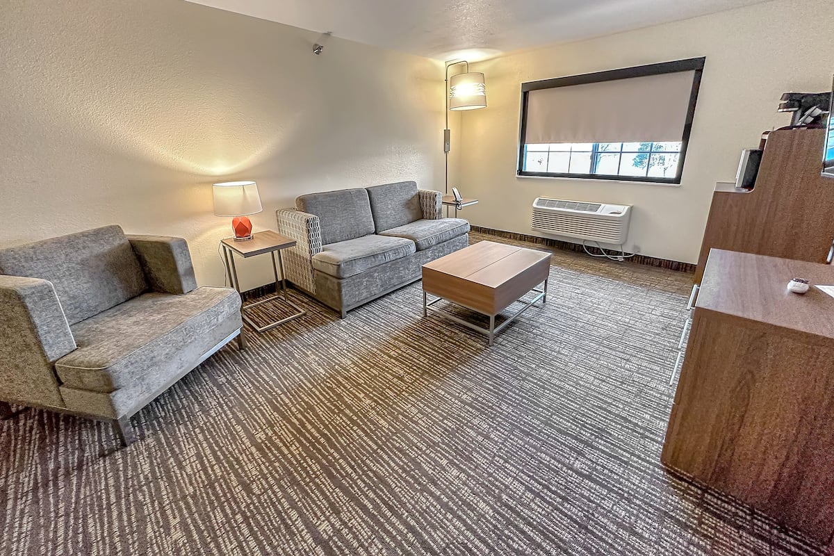 Een moderne hotelkamer met een grijze bank, een klein bijzettafeltje met een lamp, een houten salontafel en een groot raam met uitzicht.
