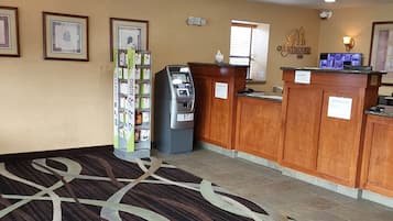 Zona de recepción de un hotel con suelo alfombrado, un quiosco de autoservicio, una caja registradora y obras de arte enmarcadas en la pared.