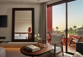 A hotel room with a large bed, a sitting area with a table and chairs, a TV mounted on the wall, and a balcony with a view of palm trees.
