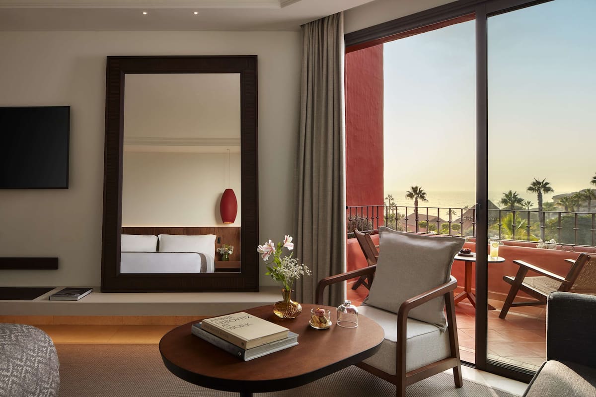 A hotel room with a large bed, a sitting area with a table and chairs, a TV mounted on the wall, and a balcony with a view of palm trees.