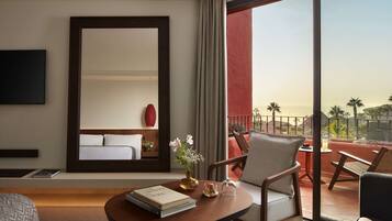 A hotel room with a large bed, a sitting area with a table and chairs, a TV mounted on the wall, and a balcony with a view of palm trees.