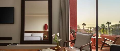 Habitación de hotel con una cama grande, zona de estar con una mesa y sillas, televisor montado en la pared y balcón con vistas a palmeras.