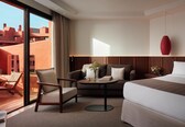 A hotel room with a bed, two armchairs, a small table, and a view of buildings through a large window.
