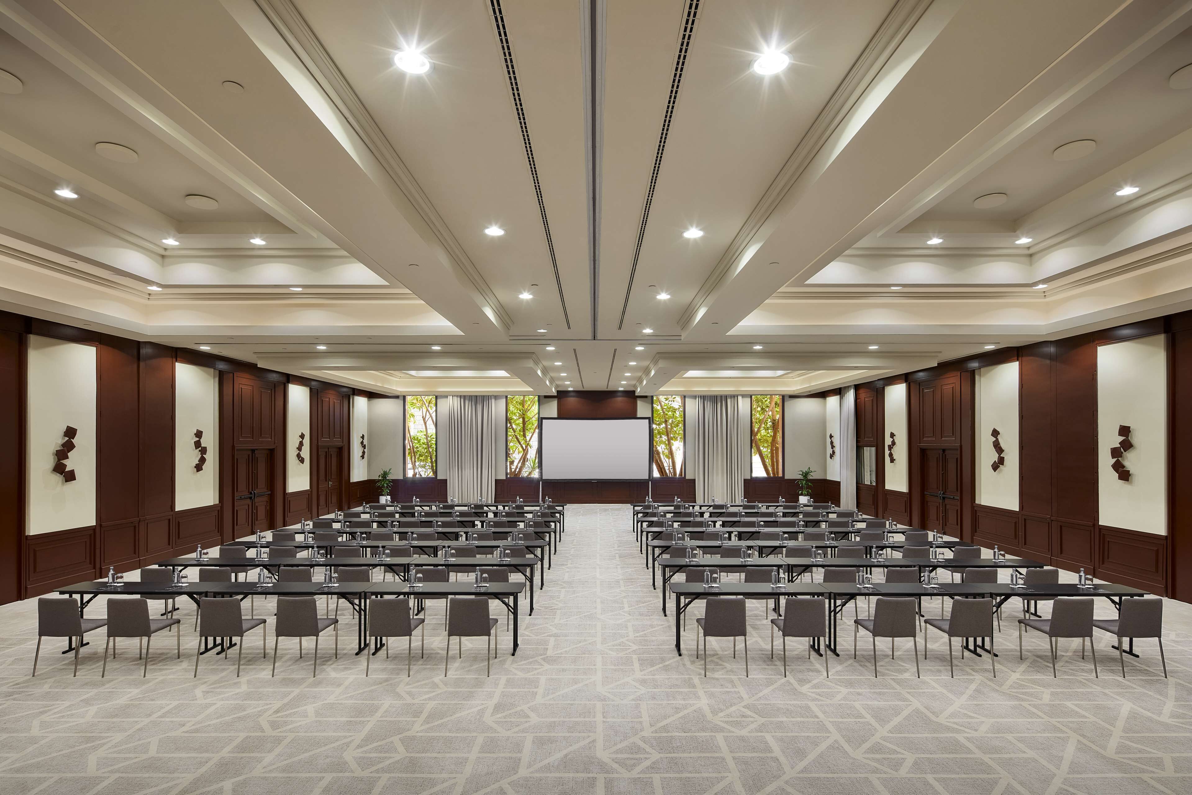 Un salón de conferencias con filas de mesas y sillas, una pantalla grande al frente y un techo alto con iluminación empotrada.