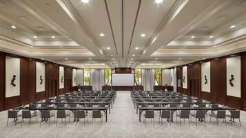 A conference room with rows of tables and chairs, a large screen at the front, and a high ceiling with recessed lighting.
