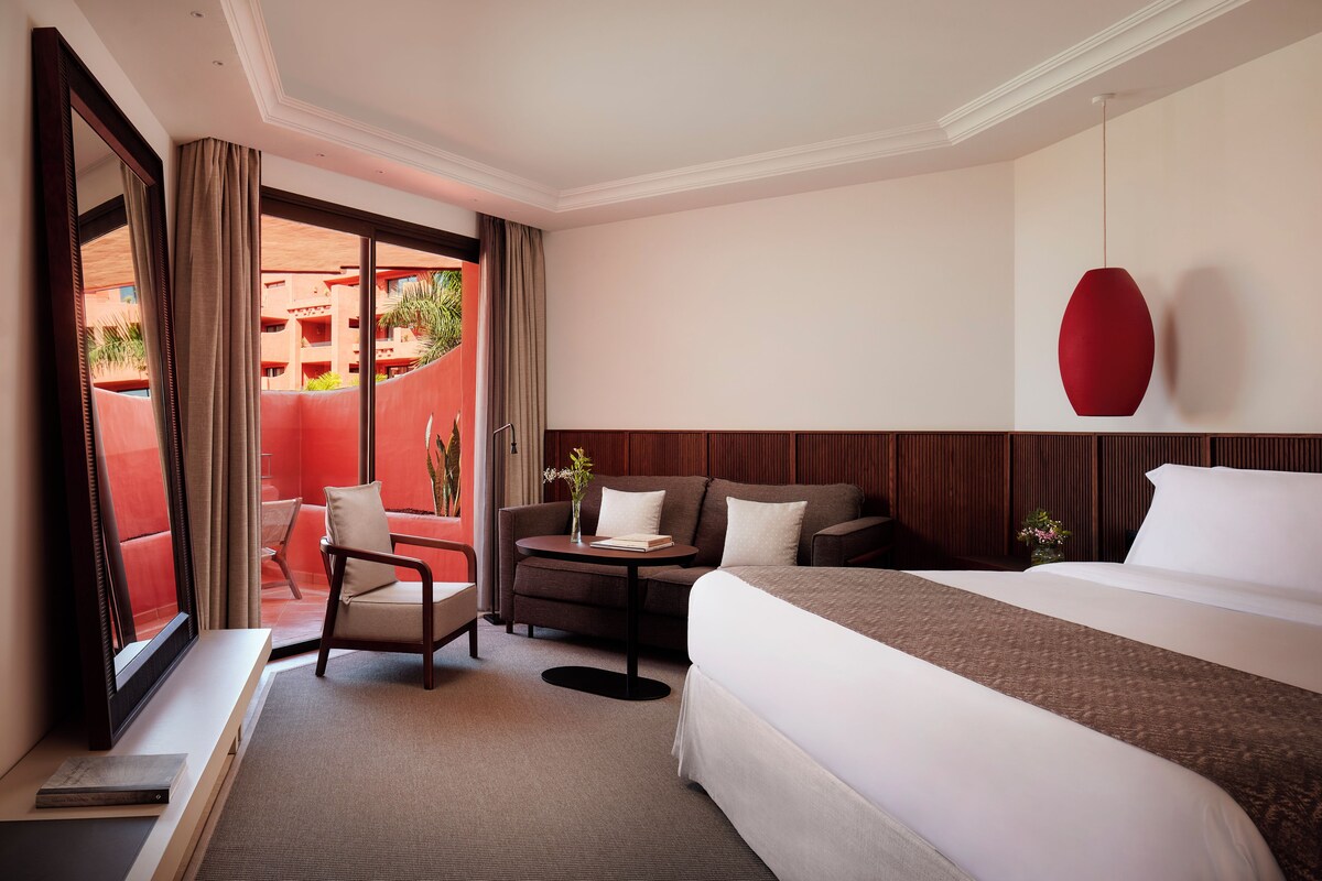 A hotel room with a large bed, a sofa, a small table, and a chair. There is a window with curtains, a red pendant light, and a view of a red building outside.