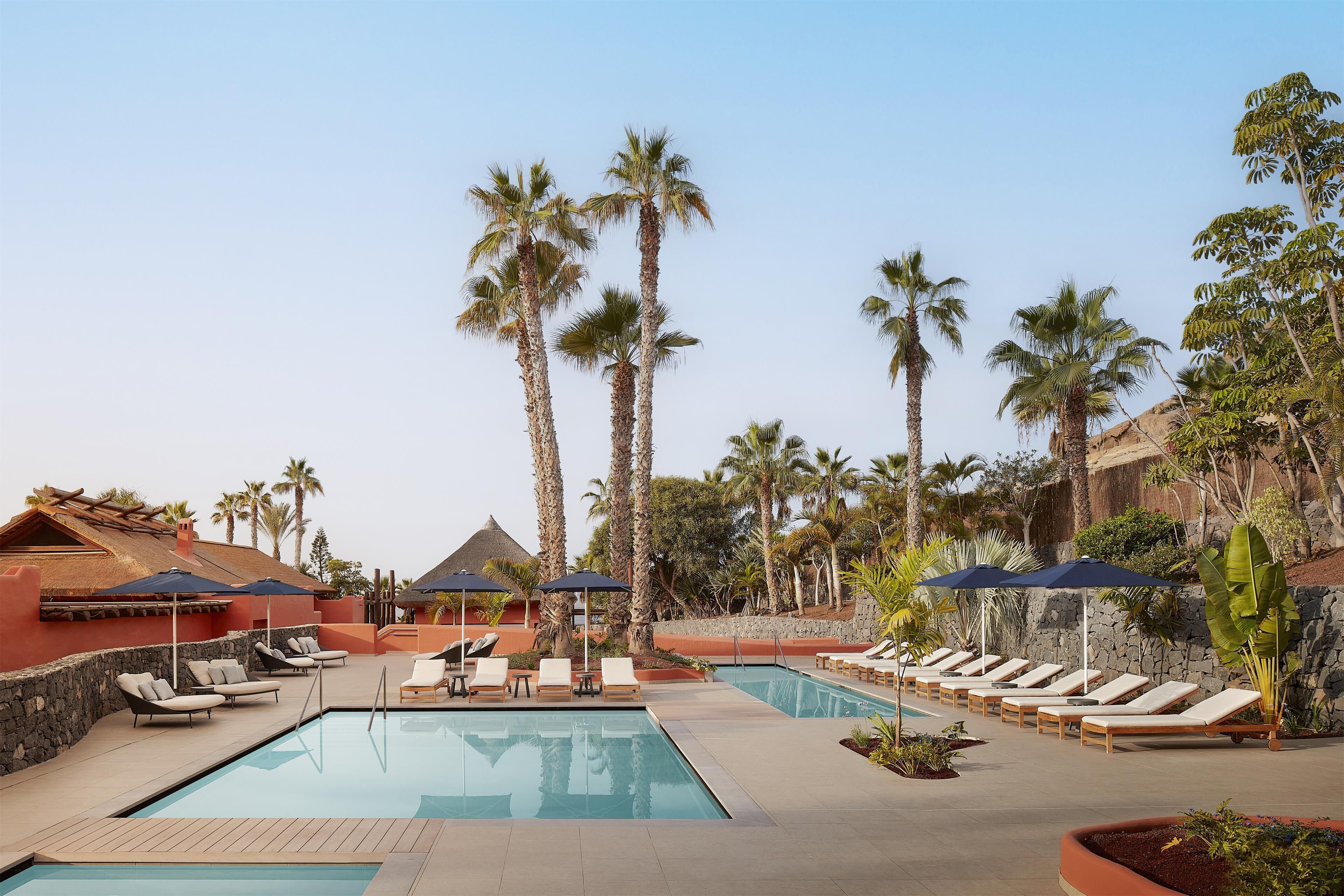A pool area with lounge chairs and umbrellas, surrounded by palm trees and a stone wall.