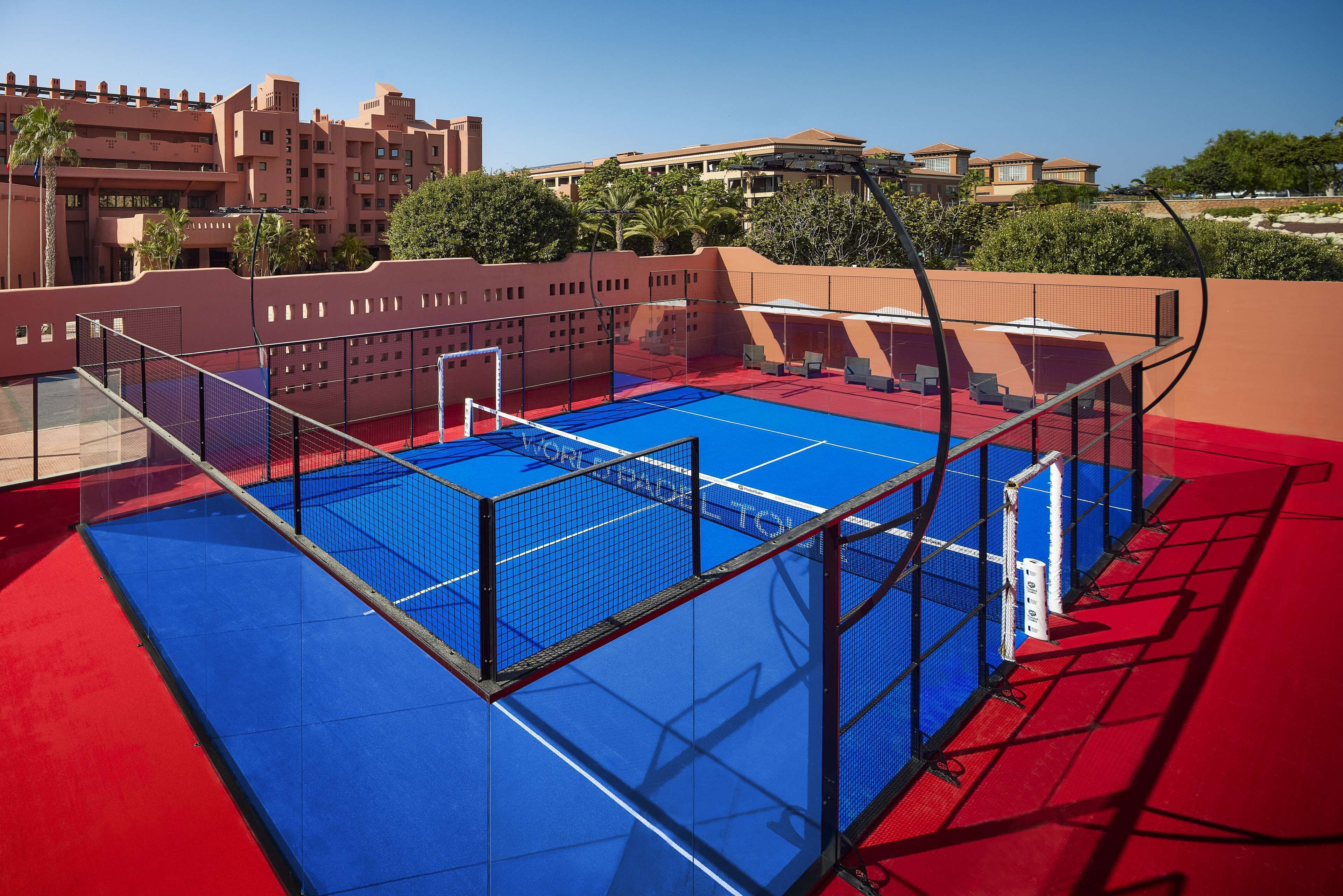 A tennis court with a net, surrounded by a red and blue floor, and a view of hotel buildings in the background.