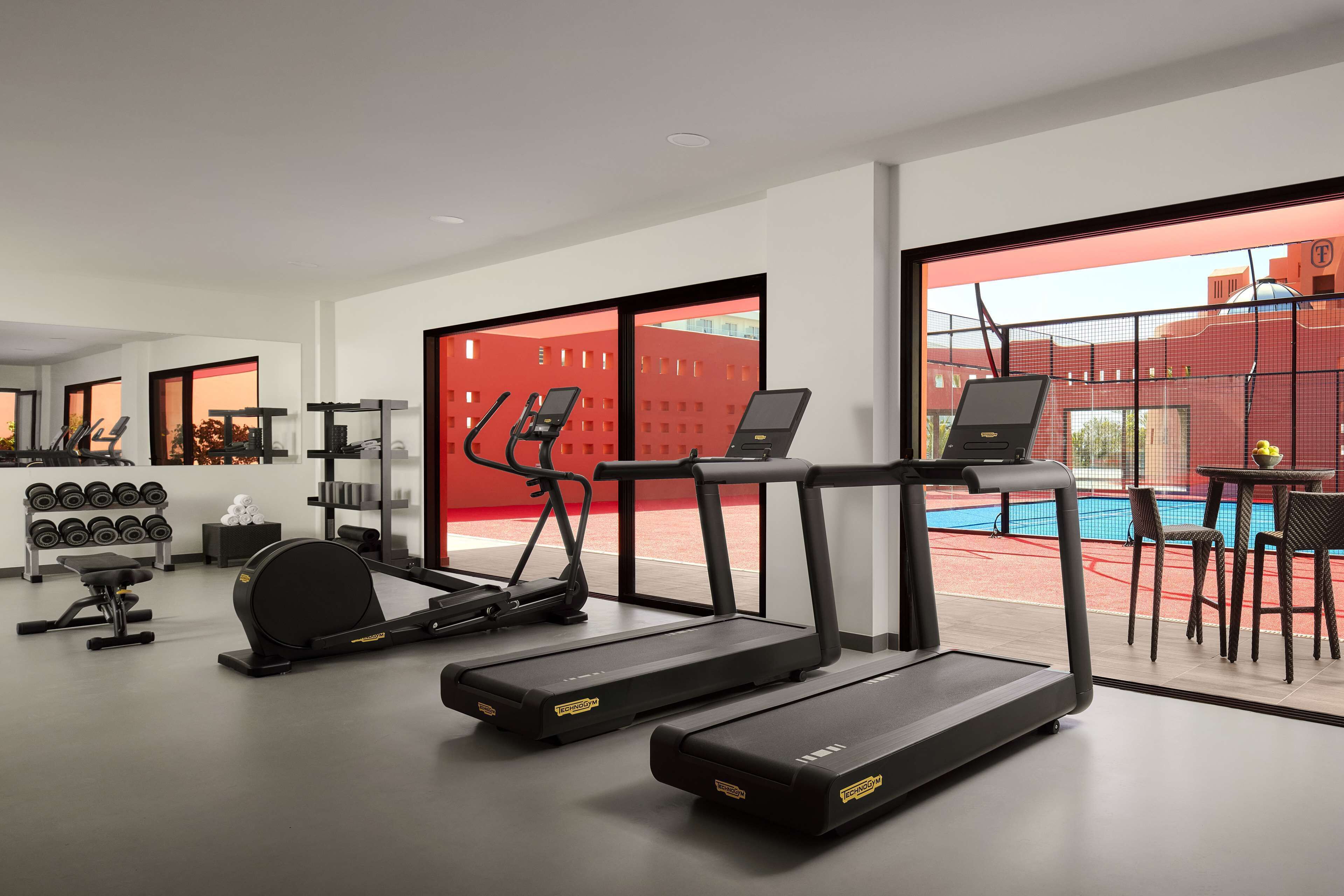 Un gimnasio moderno con cintas de correr, una máquina de remo, mancuernas y un banco. Se ve una piscina a través de amplios ventanales.