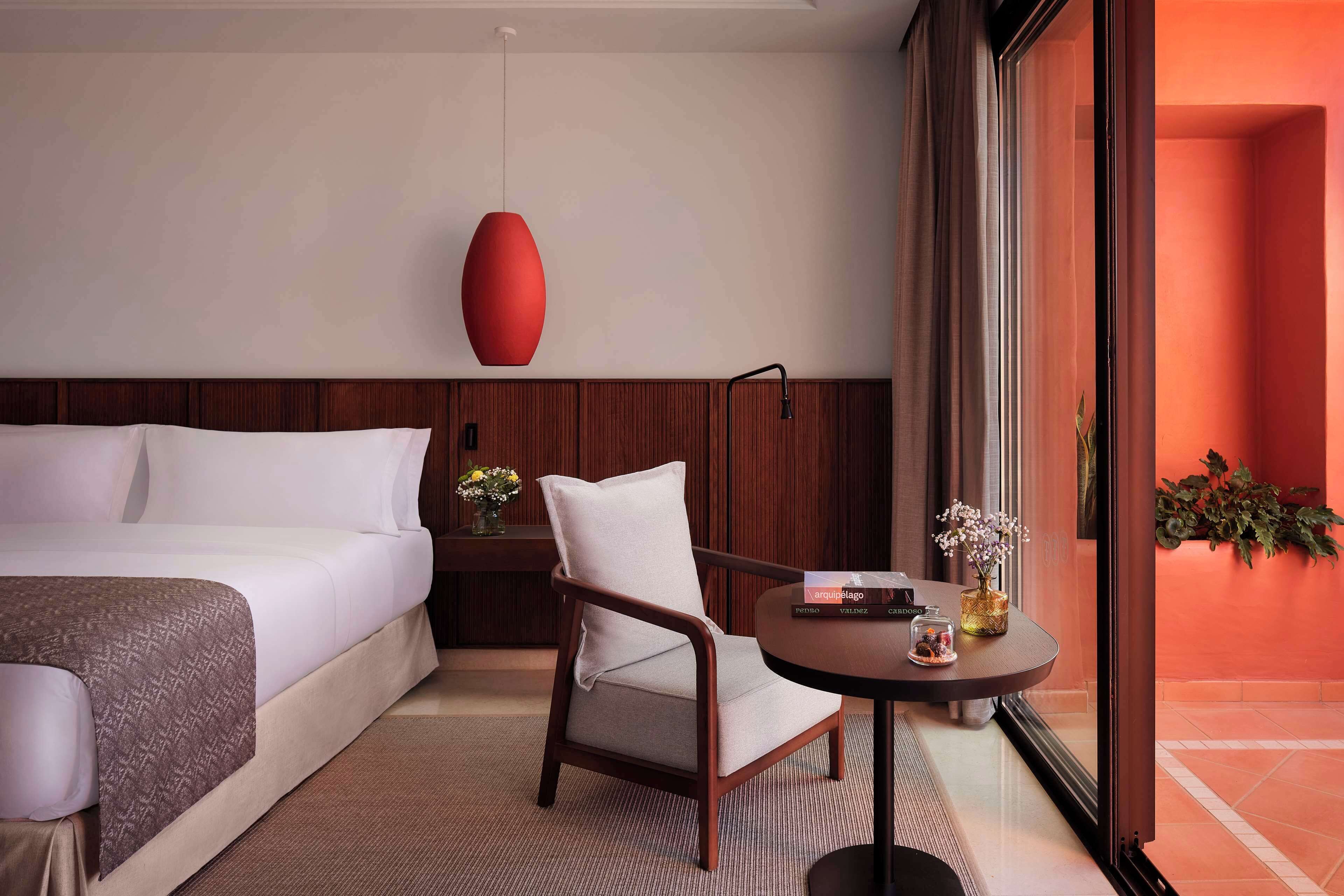 Una habitación de hotel con cama, una silla, una mesita con un florero y un ventanal grande.