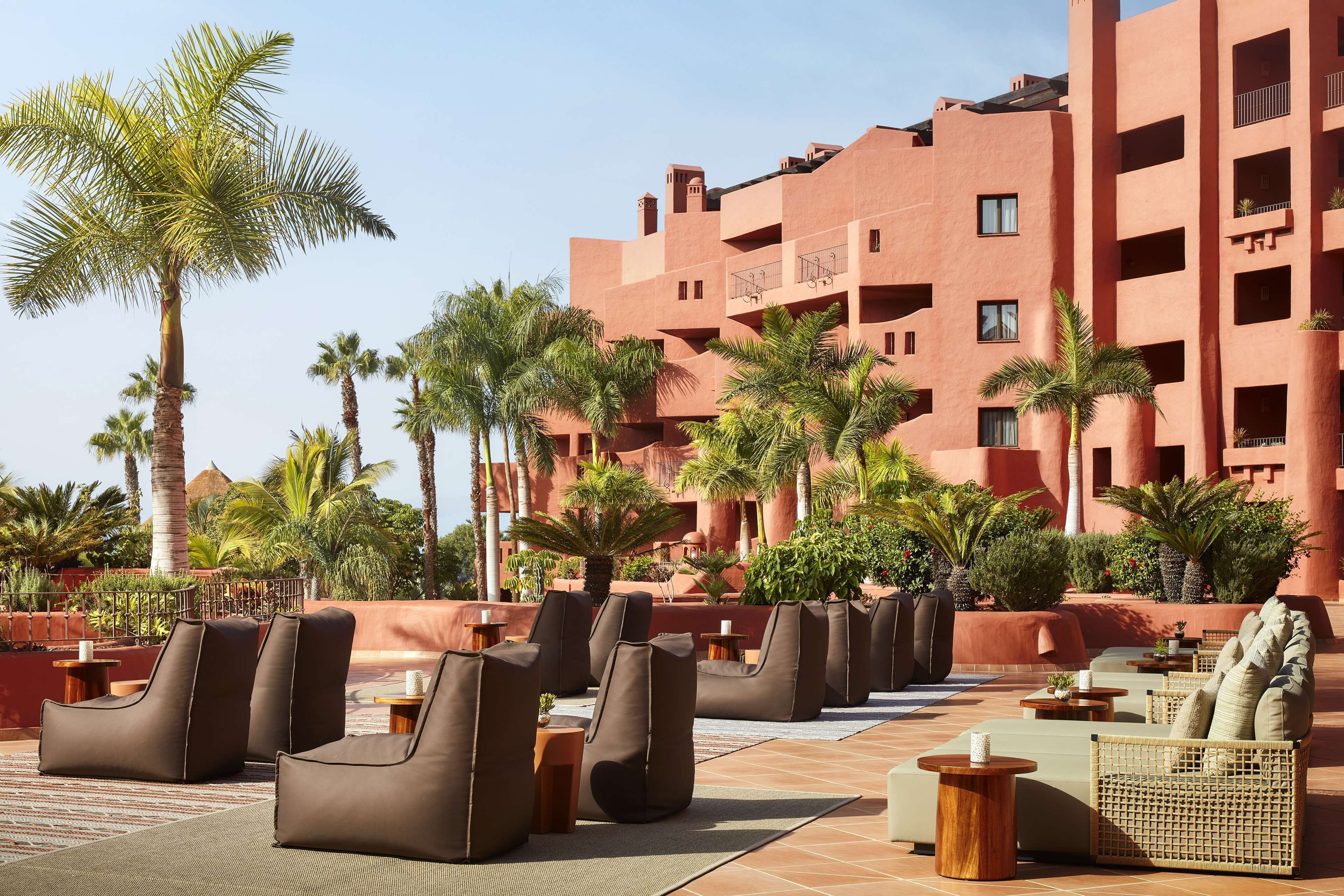 A hotel courtyard with outdoor seating, palm trees, and a red building in the background.