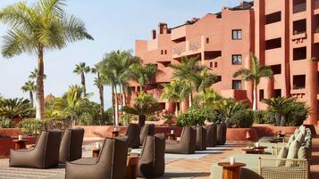 A hotel courtyard with outdoor seating, palm trees, and a red building in the background.