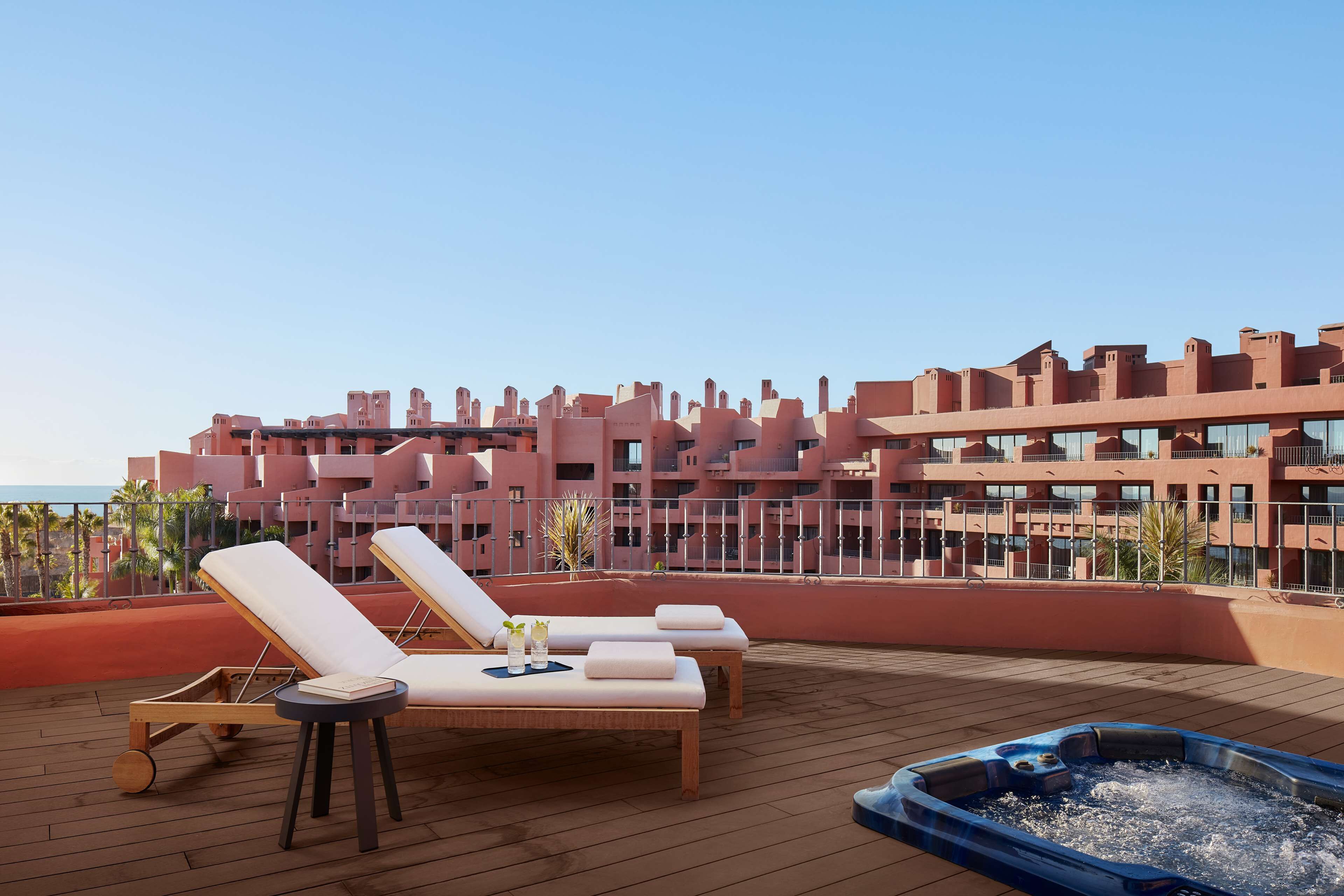 A rooftop terrace with lounge chairs and a hot tub, overlooking a large building with balconies.
