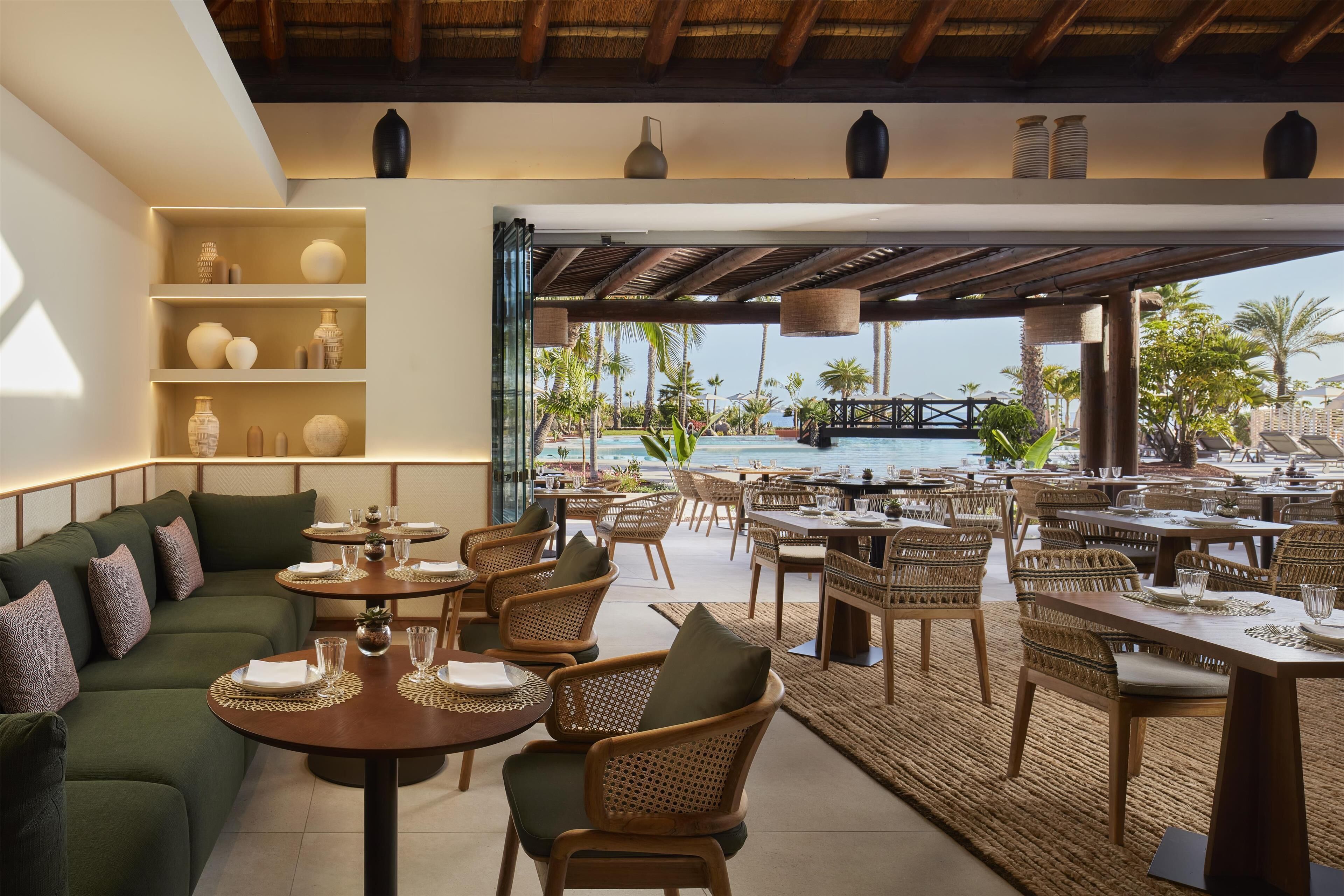 A modern outdoor dining area with round tables and wicker chairs, overlooking a pool and palm trees.