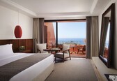 A hotel room with a bed, a desk, two chairs, a large window with a view of the sea, and a balcony with a table and chairs.