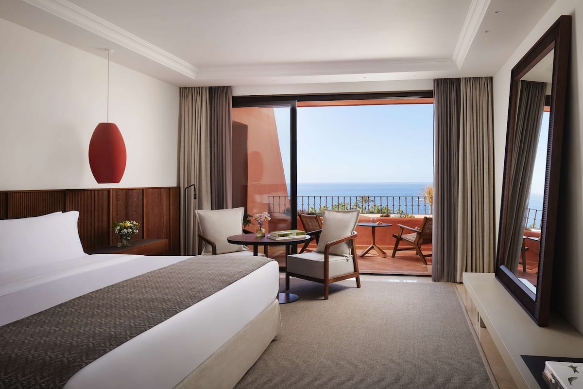 A hotel room with a bed, a desk, two chairs, a large window with a view of the sea, and a balcony with a table and chairs.
